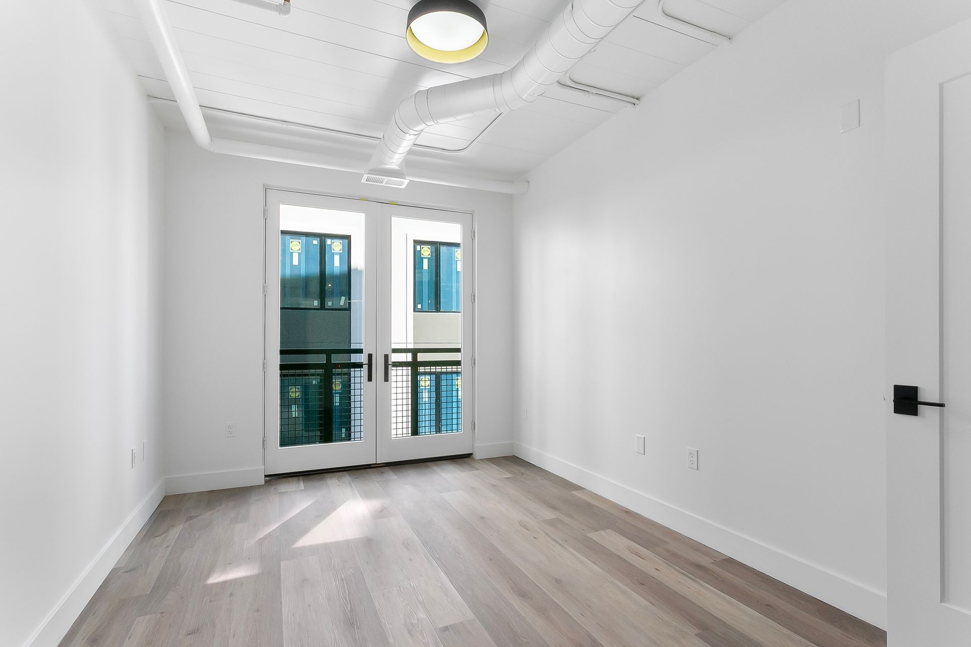 Empty white-walled room with wooden floor, French doors to a balcony, and a ceiling light.