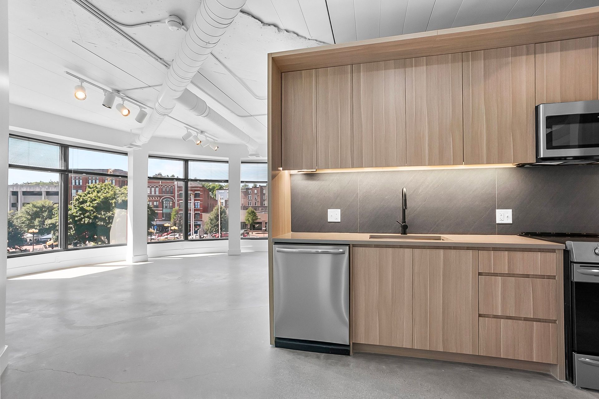 Modern kitchen with light wood cabinetry, stainless steel appliances, and large windows overlooking a city street.