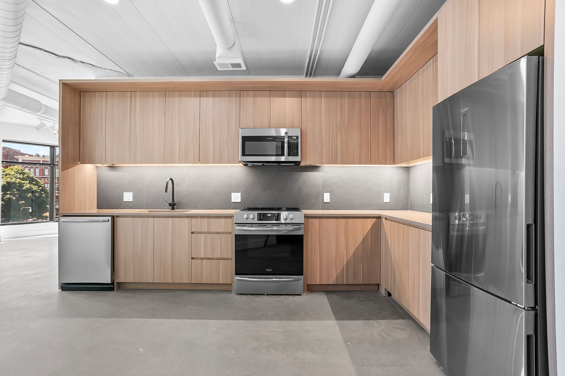 Modern kitchen with light wood cabinets, stainless steel appliances, and a gray concrete floor.