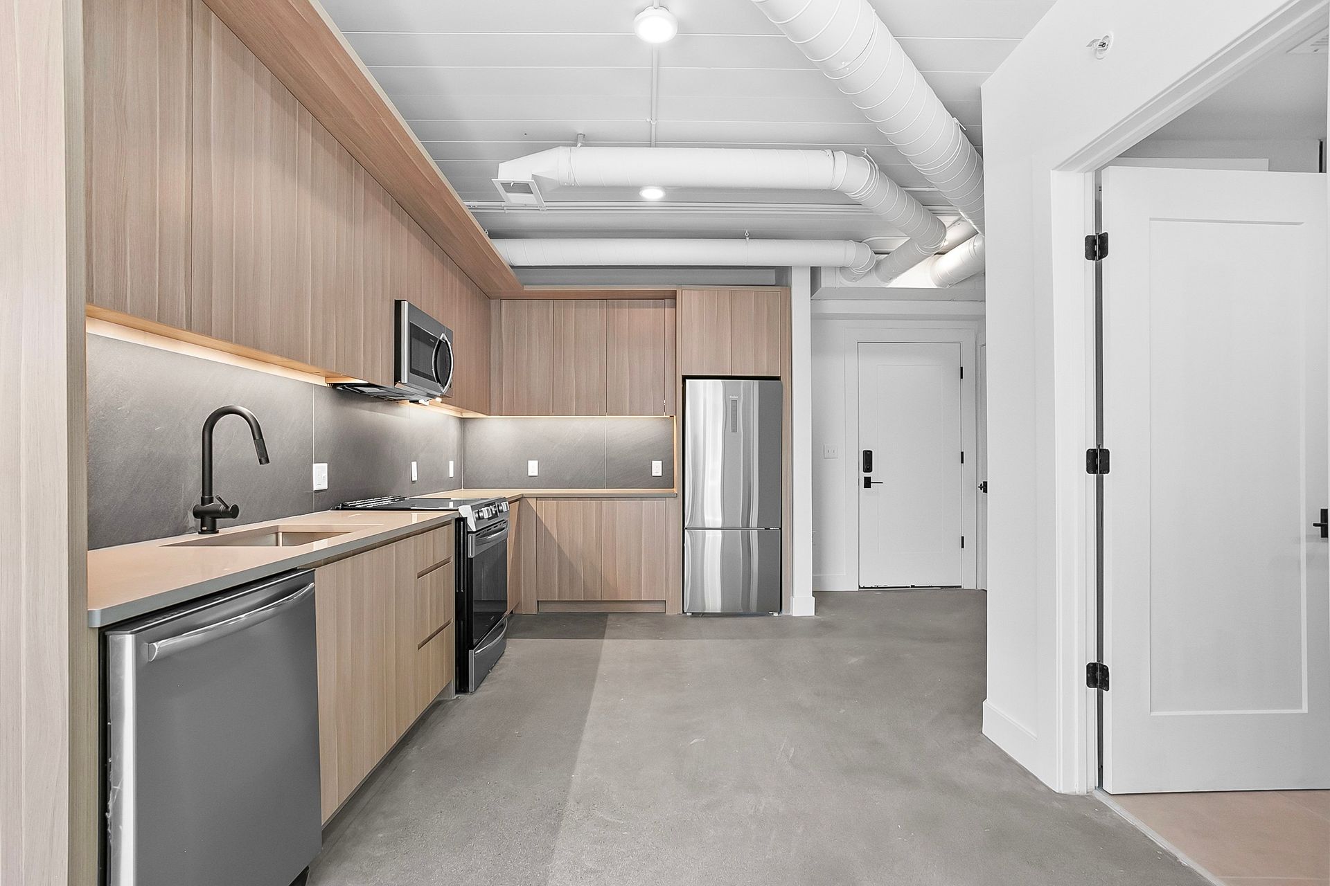 Modern kitchen with light wood cabinets, stainless steel appliances, and concrete flooring.