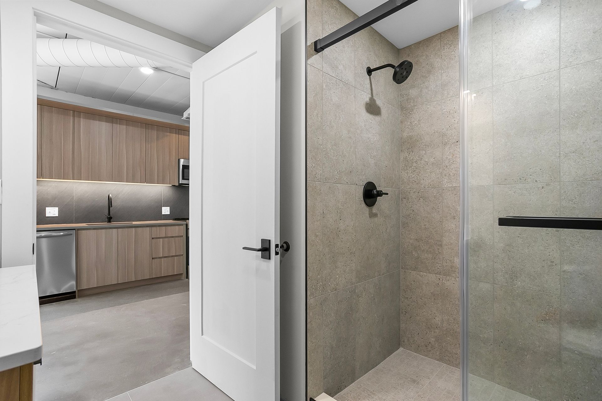 Bathroom with shower and open white door leading to a kitchen with wooden cabinets and a stainless steel refrigerator.