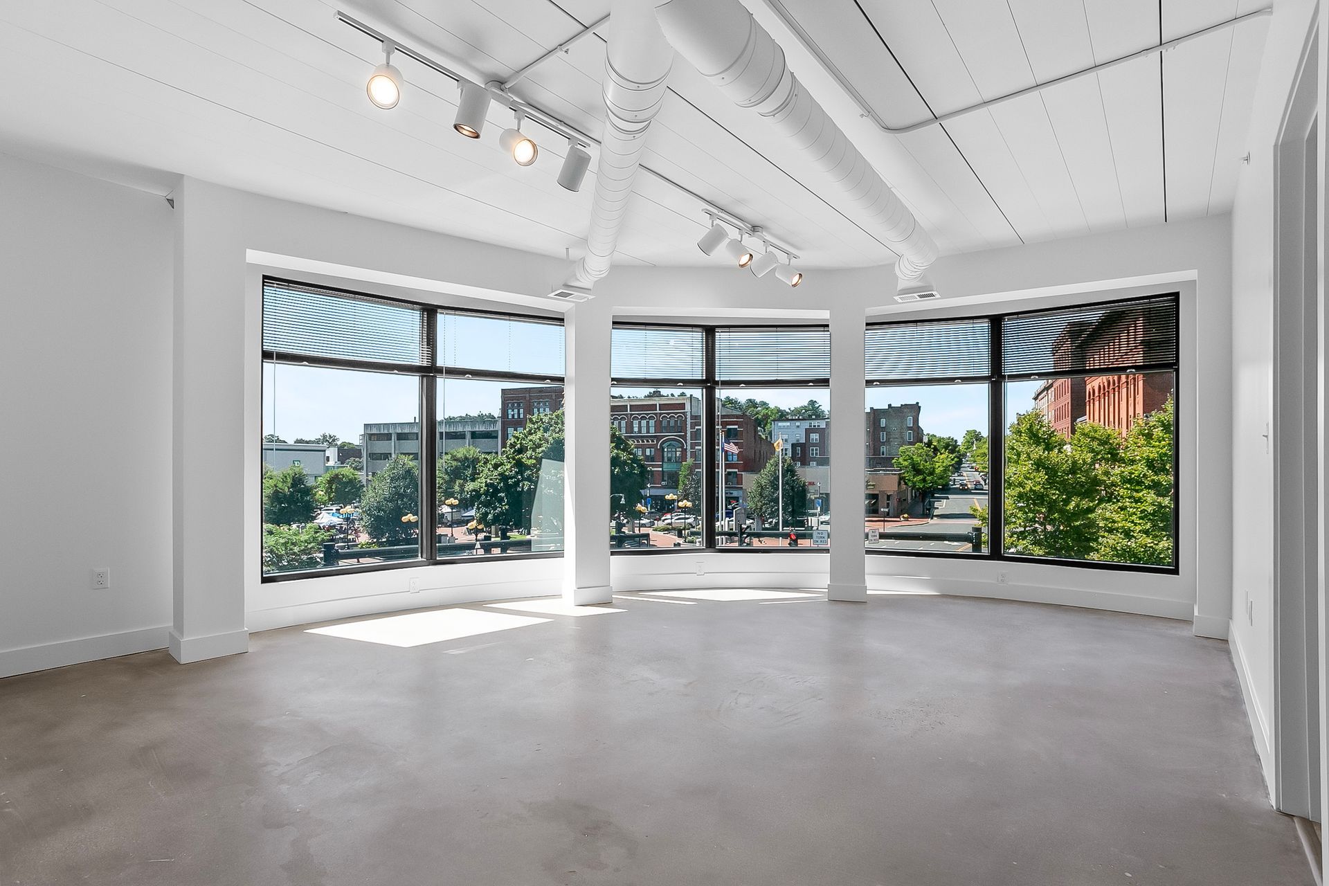 Empty white-walled room with large curved windows overlooking a city street. Track lighting on the ceiling.