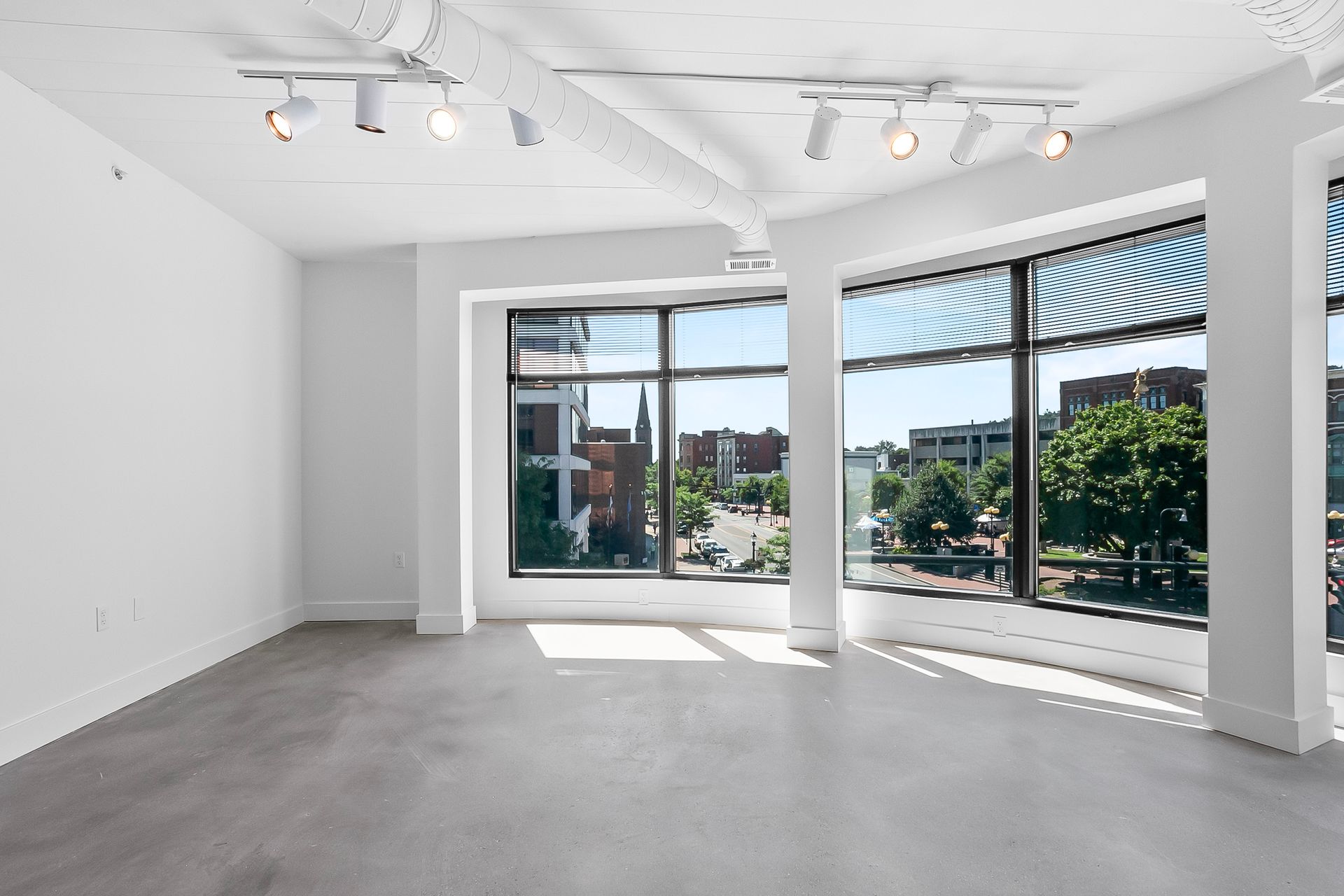 Empty, modern, bright room with large windows overlooking a city street. Grey floor, white walls and ceiling, track lighting.