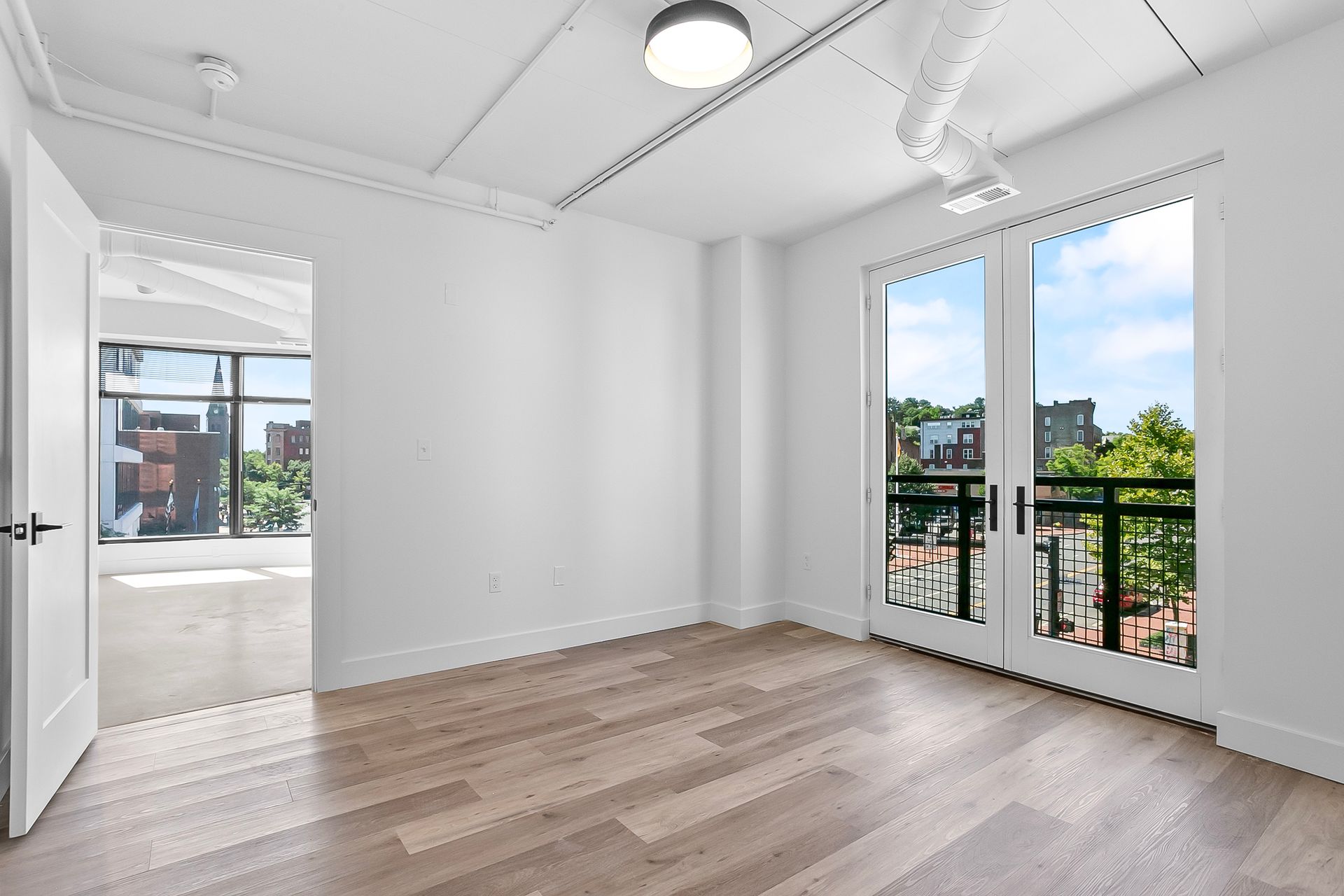 Empty room with light wood floors, white walls, and French doors to a balcony overlooking a city.