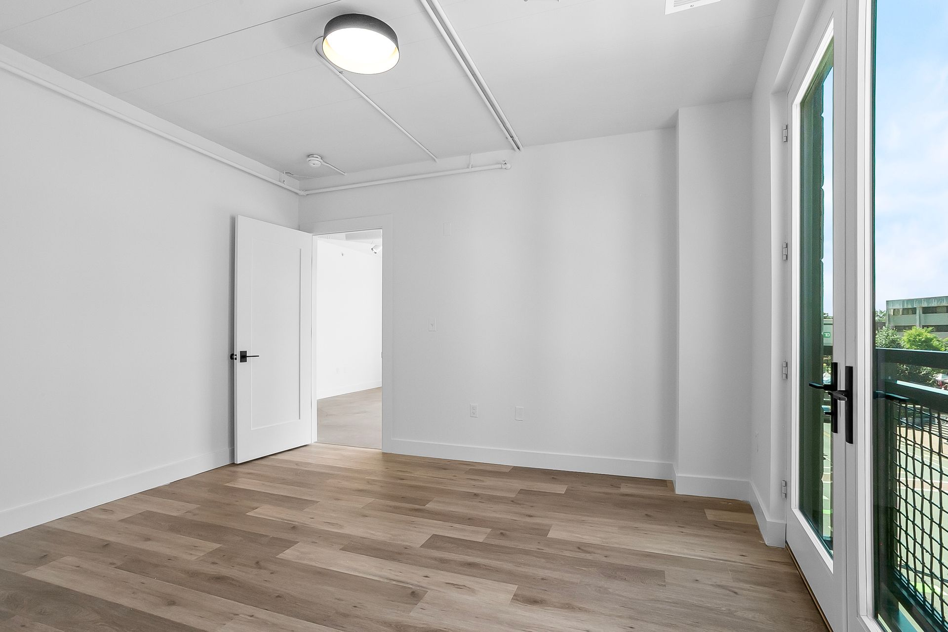 Empty, light-filled room with wood flooring, white walls, and a door leading to a balcony.