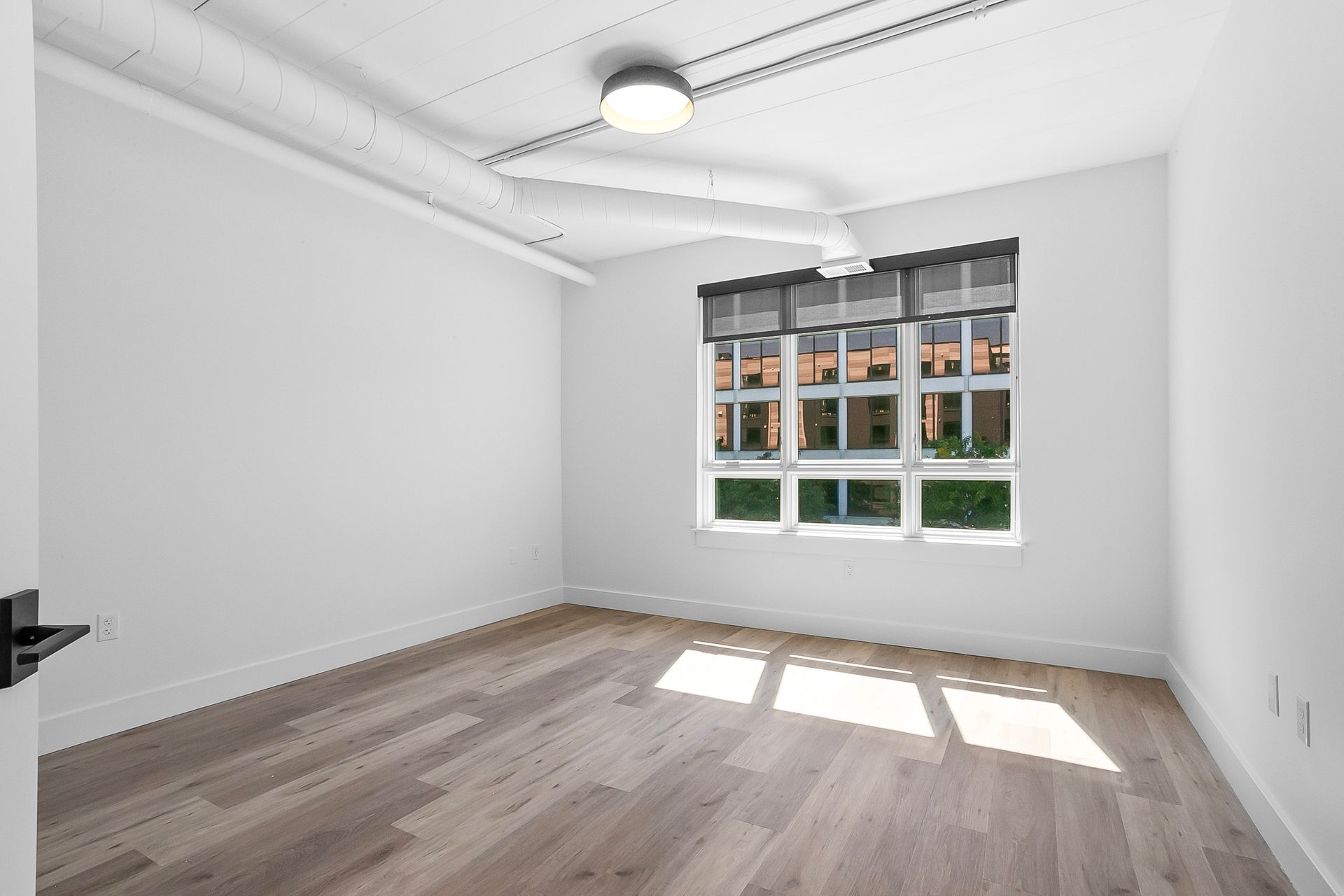 Empty room with wooden floors, large window, white walls, and industrial-style ceiling elements.