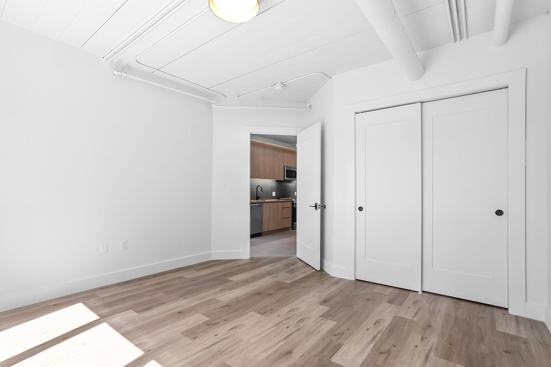 Empty white-walled room with hardwood floors, a closet, and an open doorway to a kitchen.