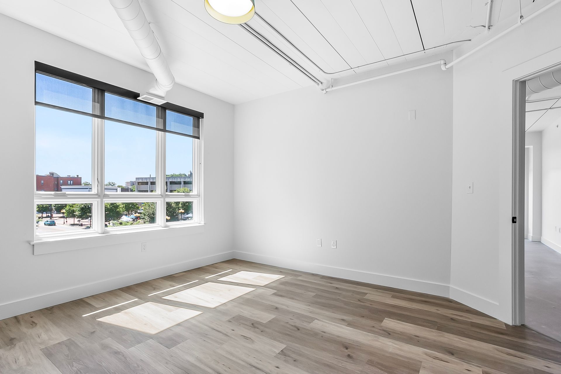 Empty, bright room with large window, light wood floor, and white walls.