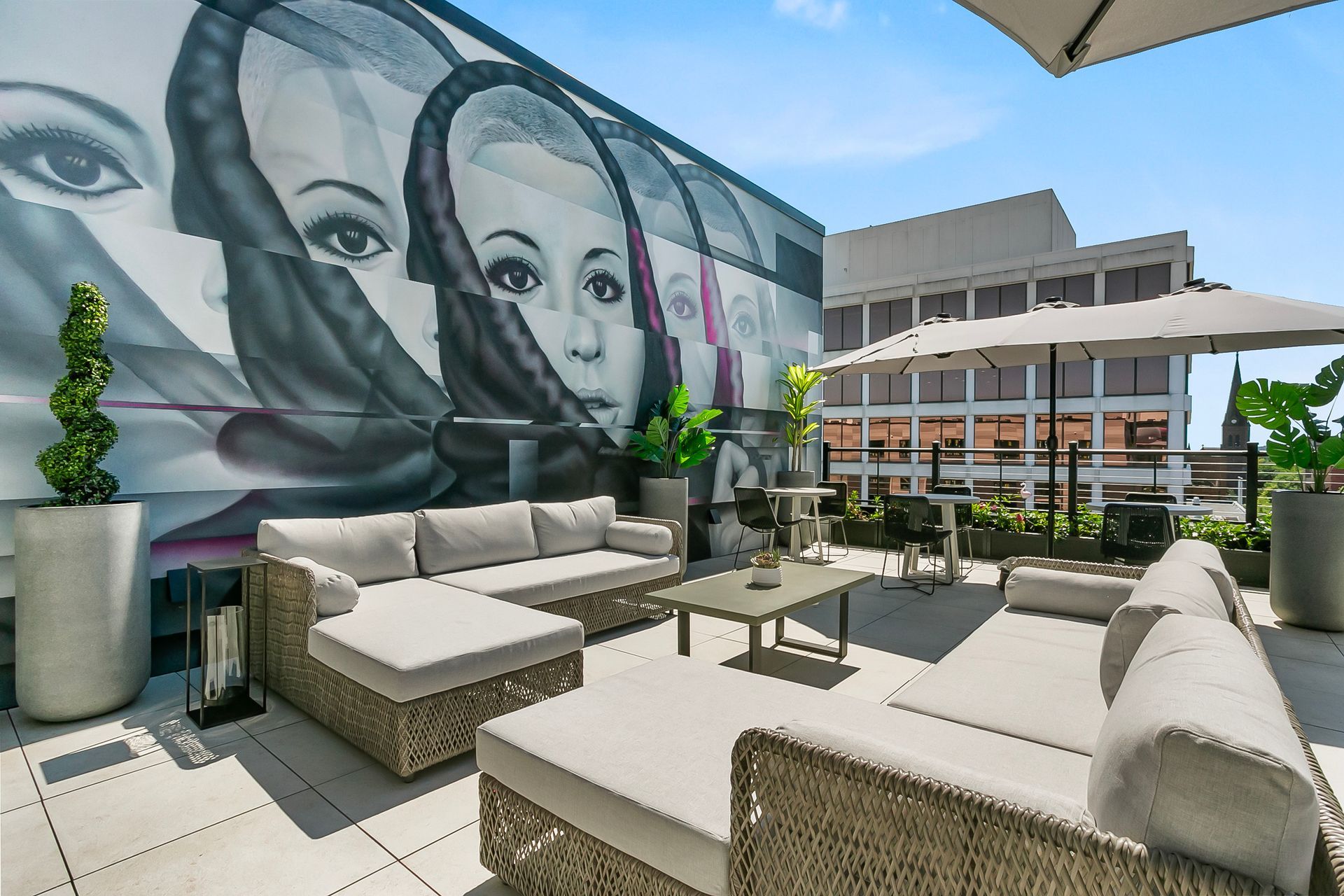Rooftop patio with gray couches, tables, and umbrella, featuring a large mural of a woman's face.