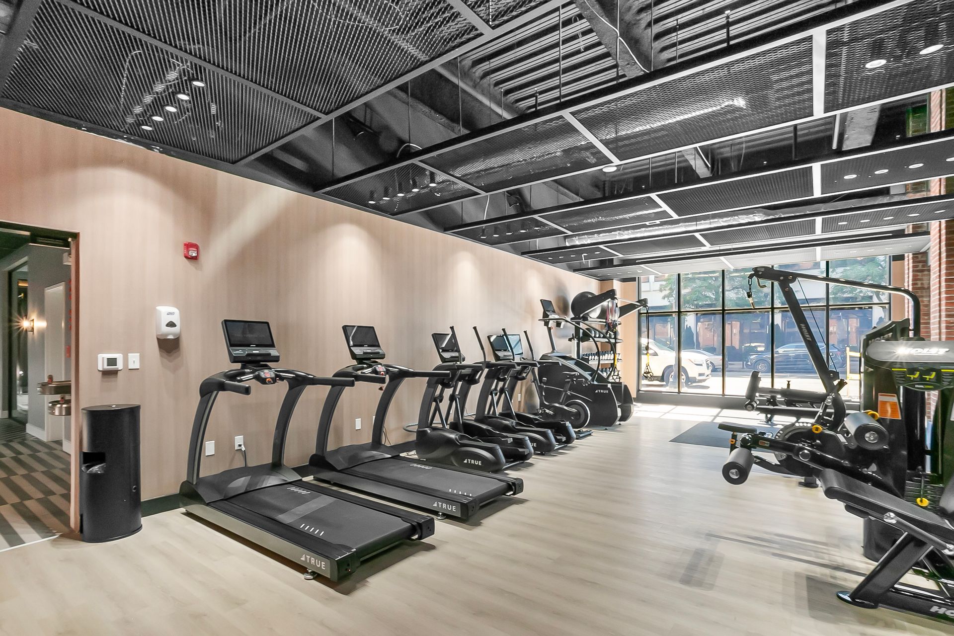 Gym interior with cardio equipment and weights, light wood floors, neutral walls, and a black ceiling.