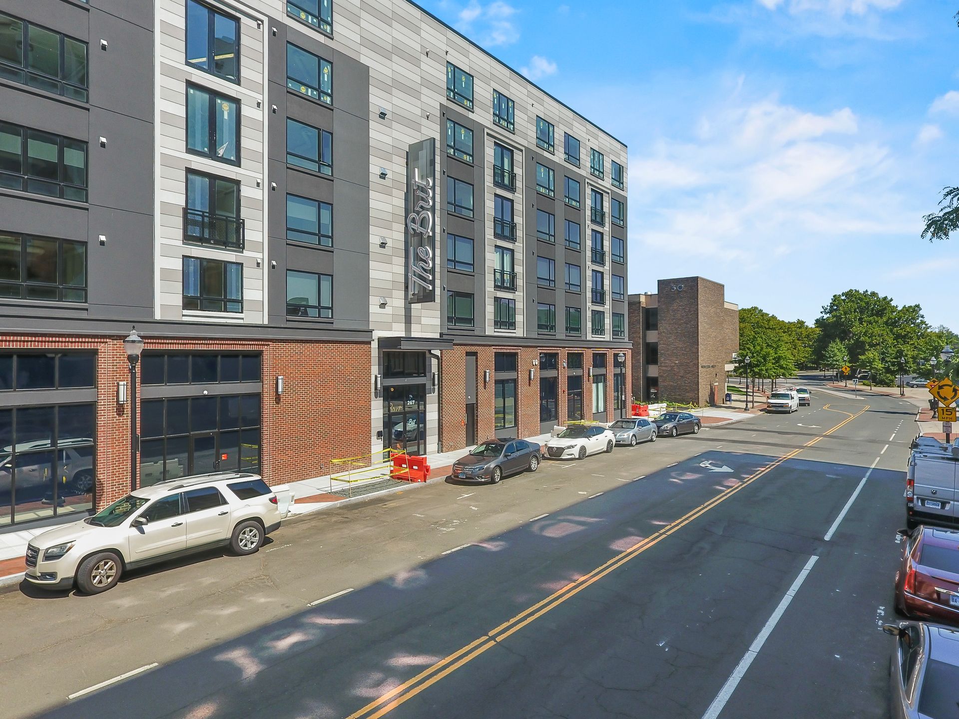 Modern apartment building with retail on street, cars parked along the road, blue sky.