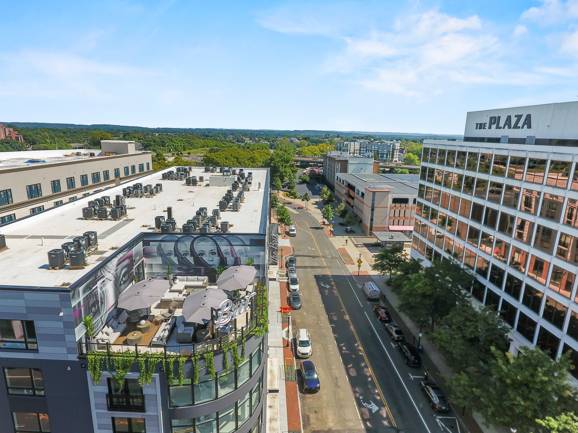 Aerial view of a city street lined with buildings and cars, with a green tree-filled skyline in the background.