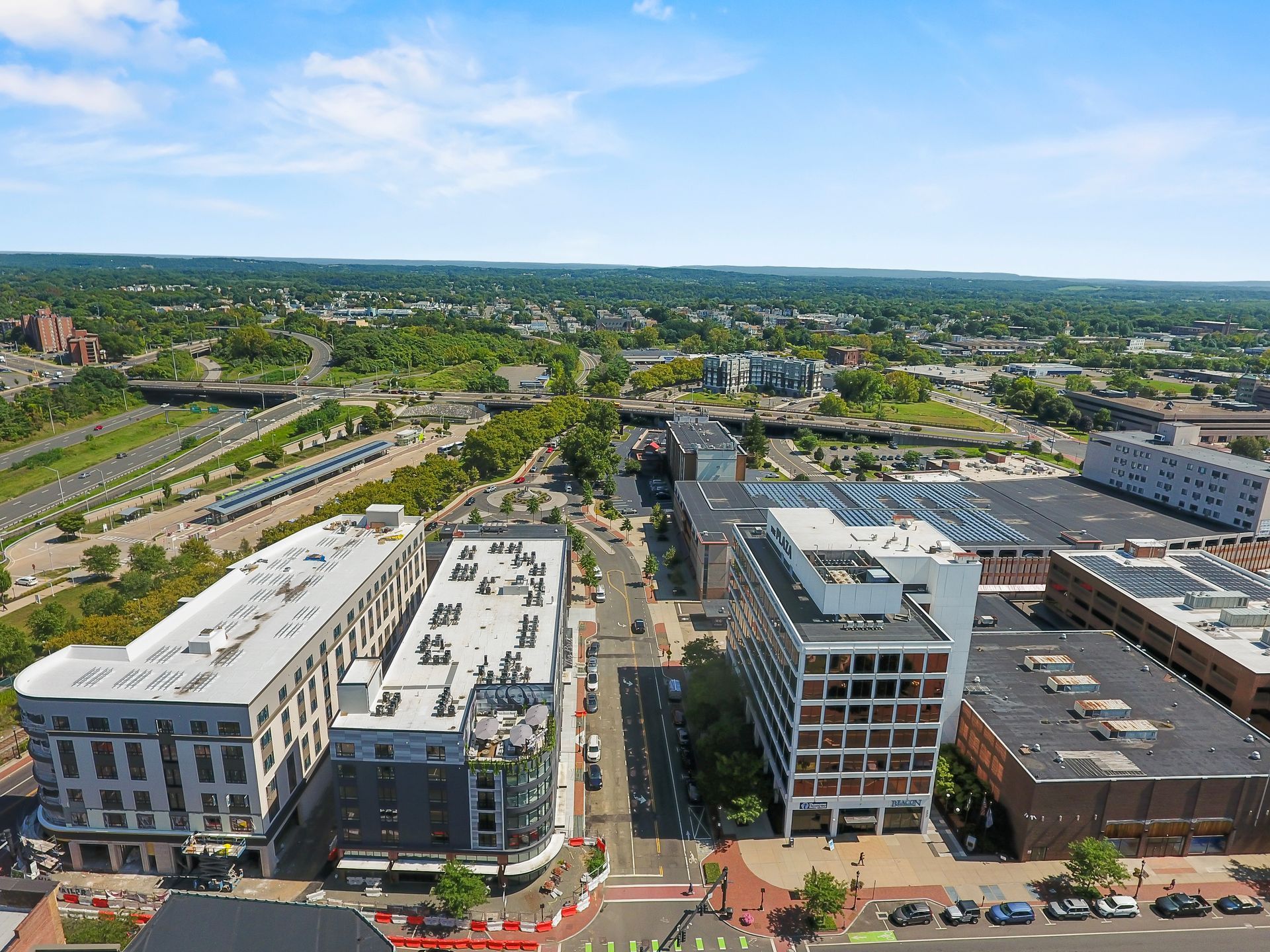 Aerial view of a city street with buildings, parked cars, and a train station in the distance under a blue sky.