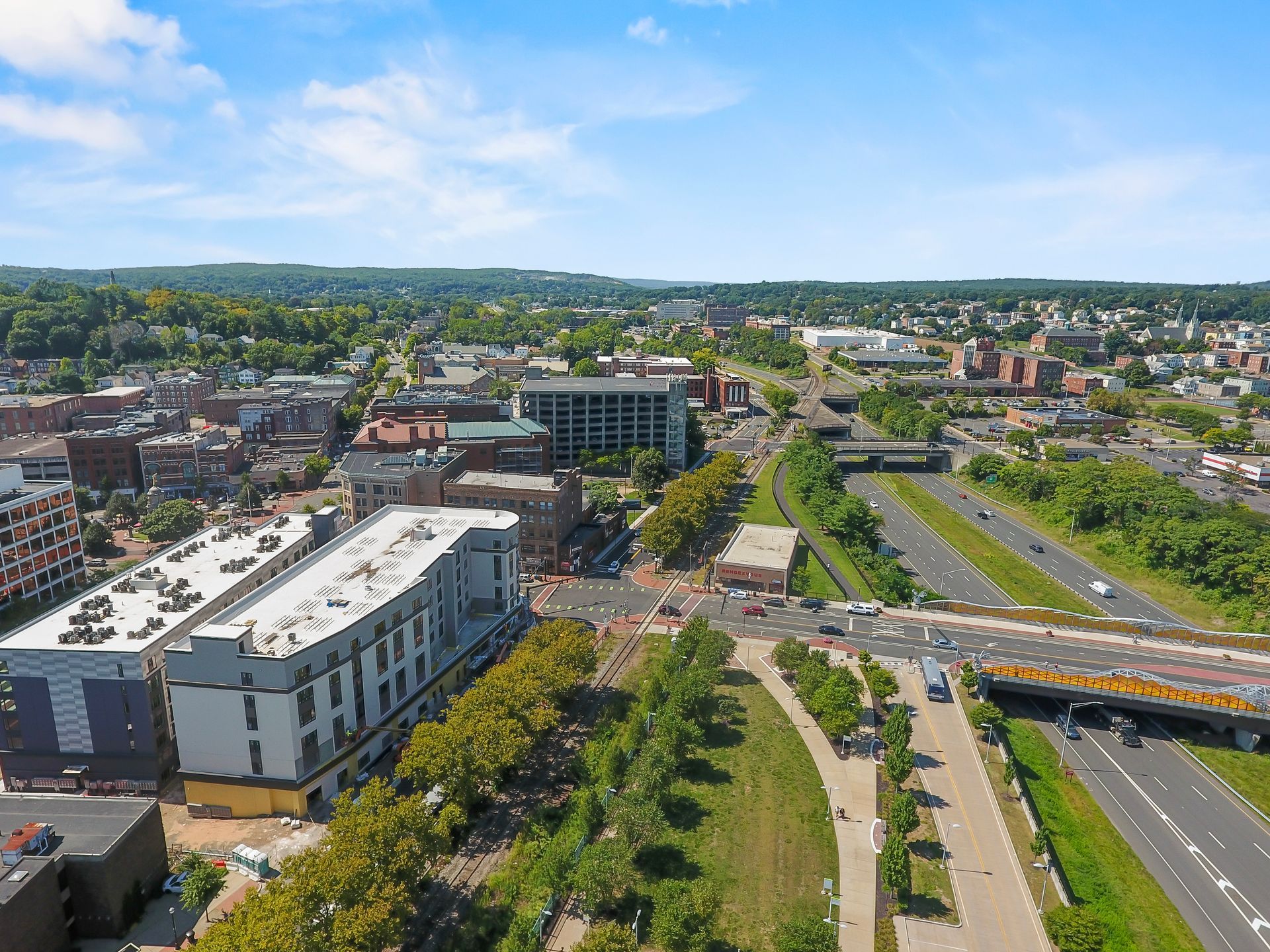 Aerial view of a city with buildings, a highway, and green spaces under a blue sky.