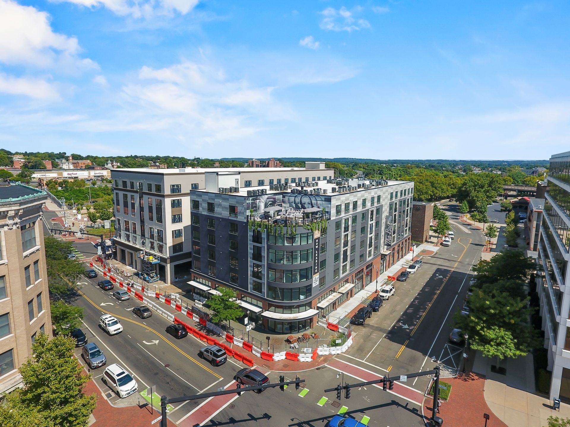 Aerial view of a multi-story building on a city street corner; construction barricades along sidewalk, cars on road, blue sky.