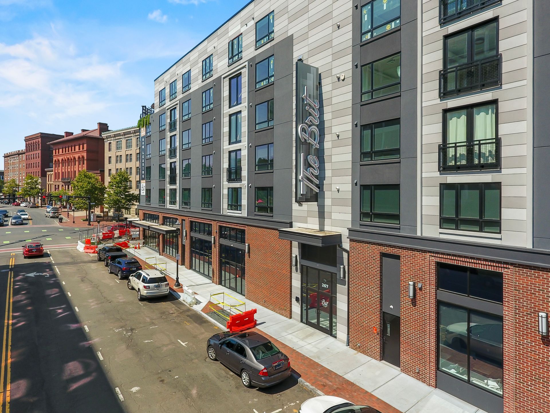 Modern apartment building on a city street, gray facade, brick accents, cars parked, sunny day.