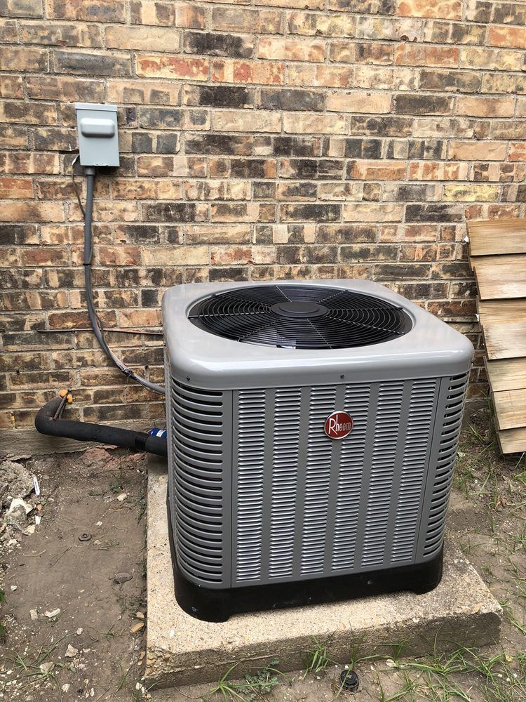 A gray air conditioner is sitting on a concrete platform in front of a brick wall.