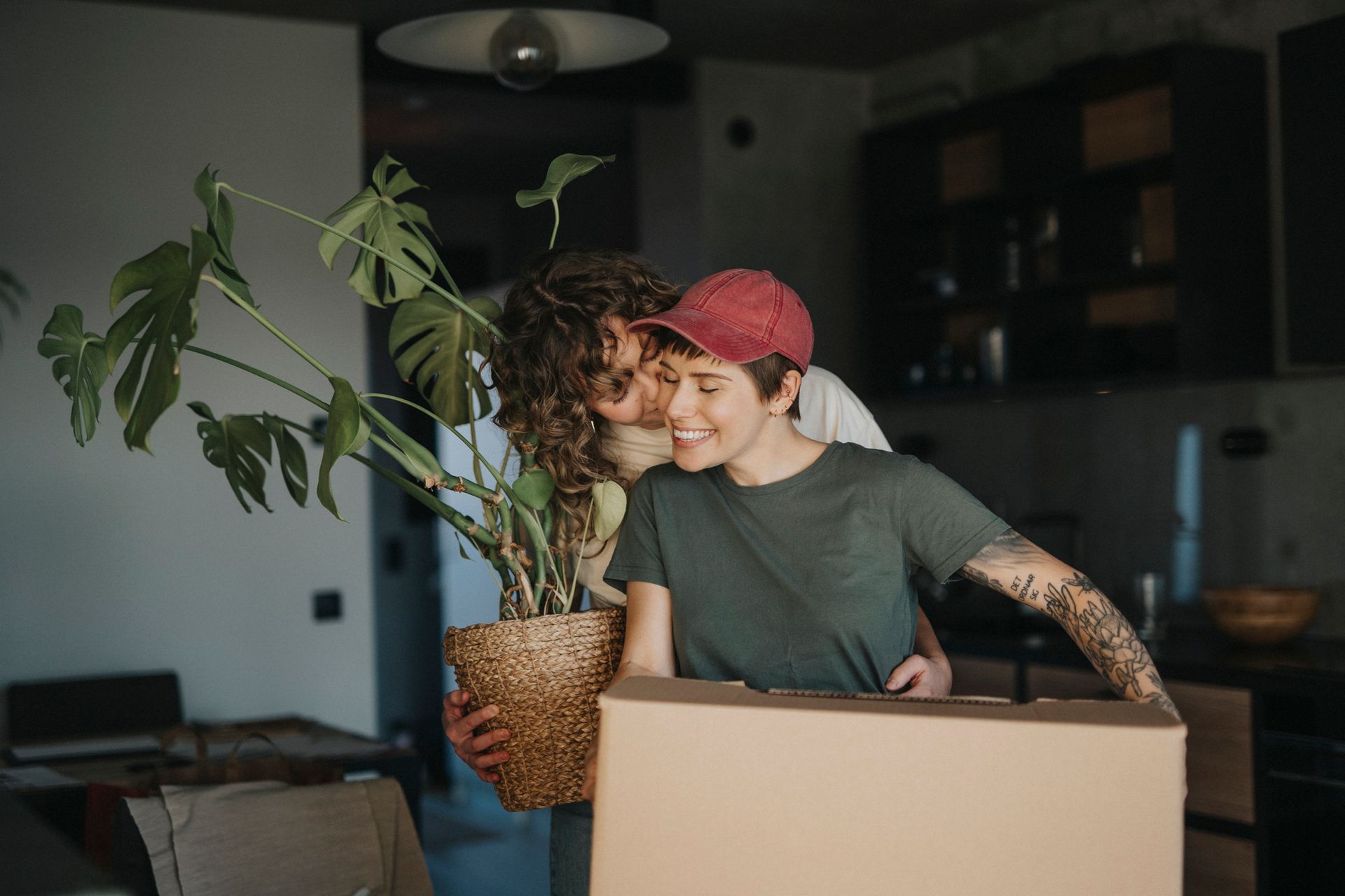 Una coppia disfa le valigie, tiene in mano una pianta in vaso e una scatola di cartone; sorride e si abbraccia in cucina.