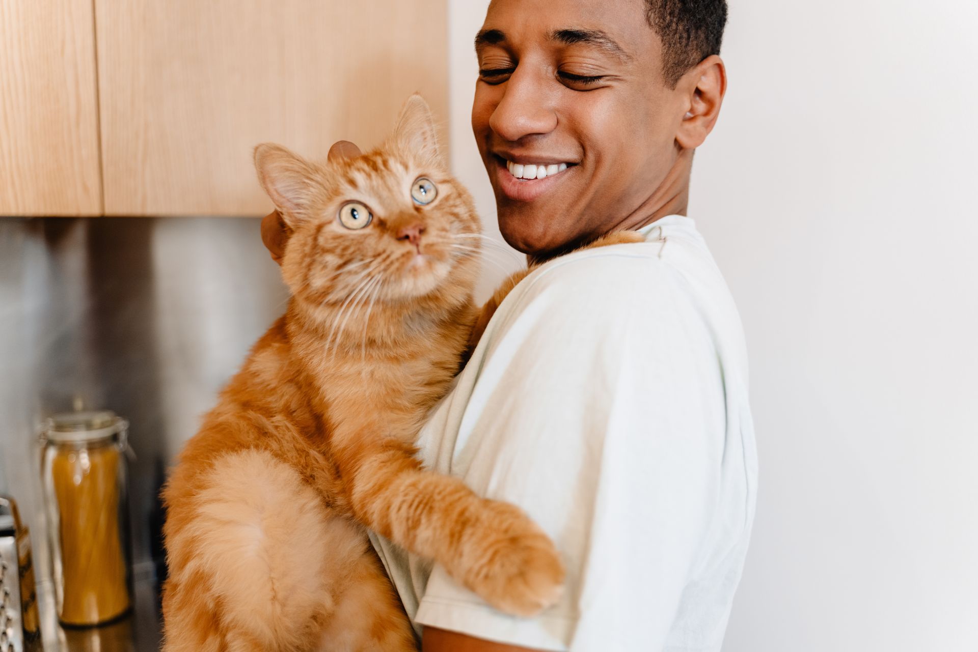 Man holding a large orange cat, both smiling in a kitchen.