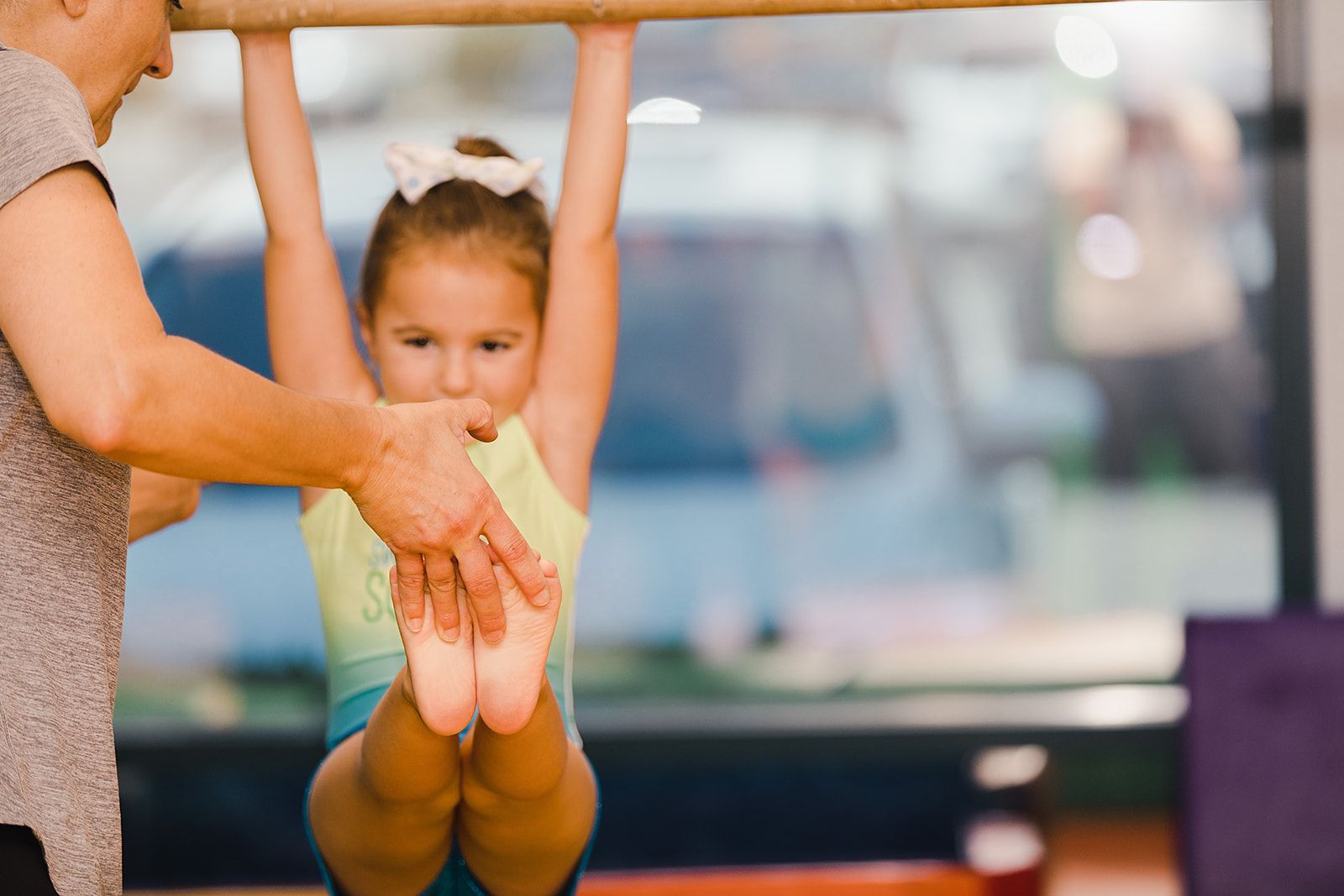 A young girl hangs from a bar, guided by an adult's hands in a dance studio.