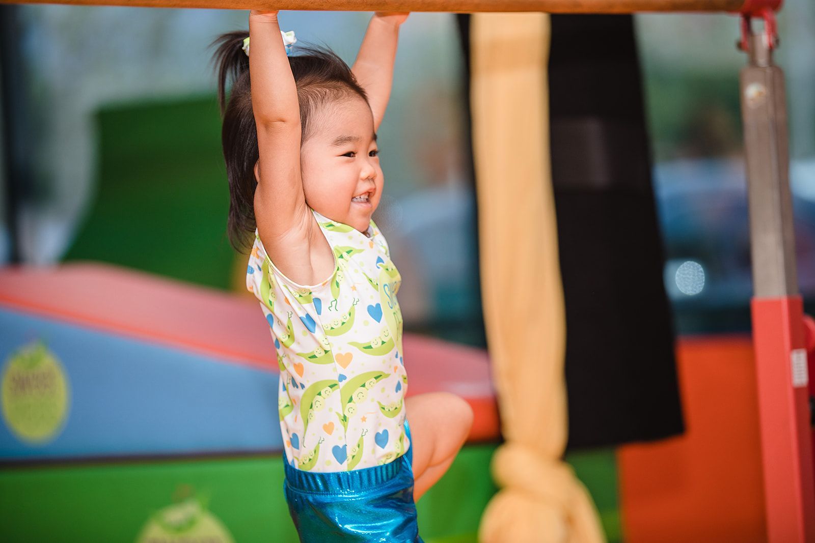 Young child smiling while hanging from a bar in a gym.