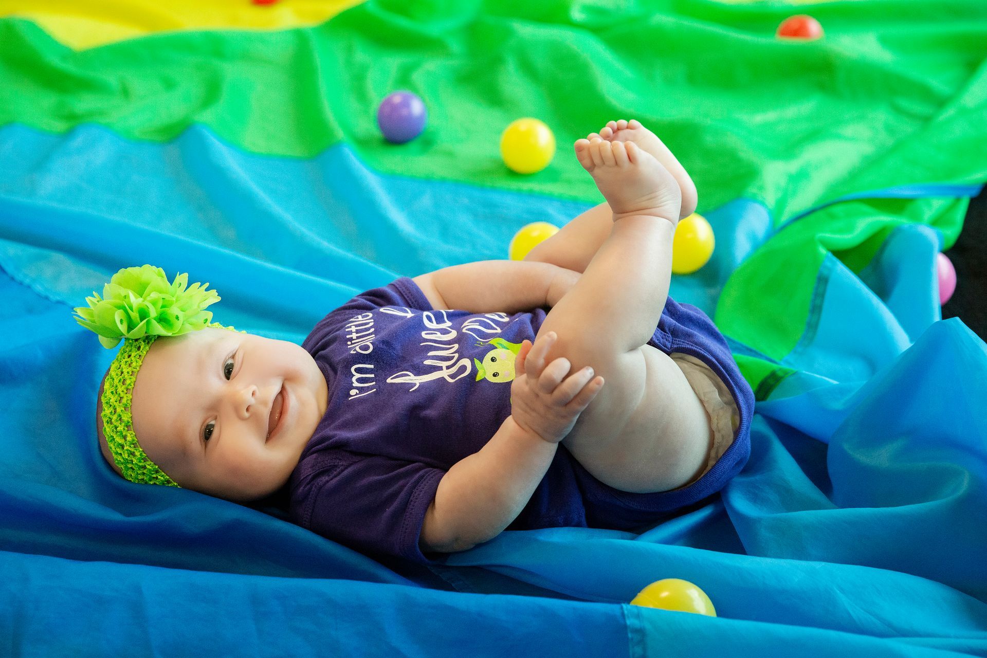 Blonde child smiles, crawling on a green surface toward the viewer in a pit filled with foam cubes.