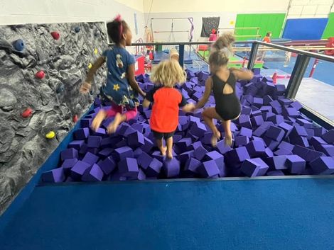 Three children jumping in a foam pit at a gymnastics gym, near a climbing wall.