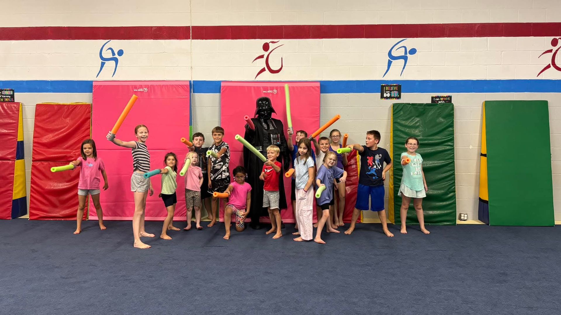 Children and Darth Vader pose with foam light sabers in a gym.