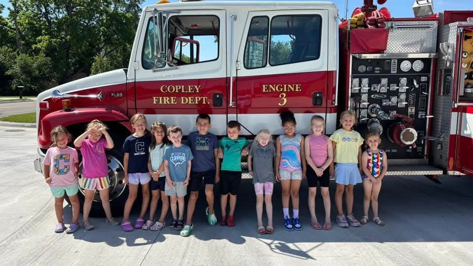 Children stand in front of a fire truck in Copley. Some smile. Sunny, outdoor setting.