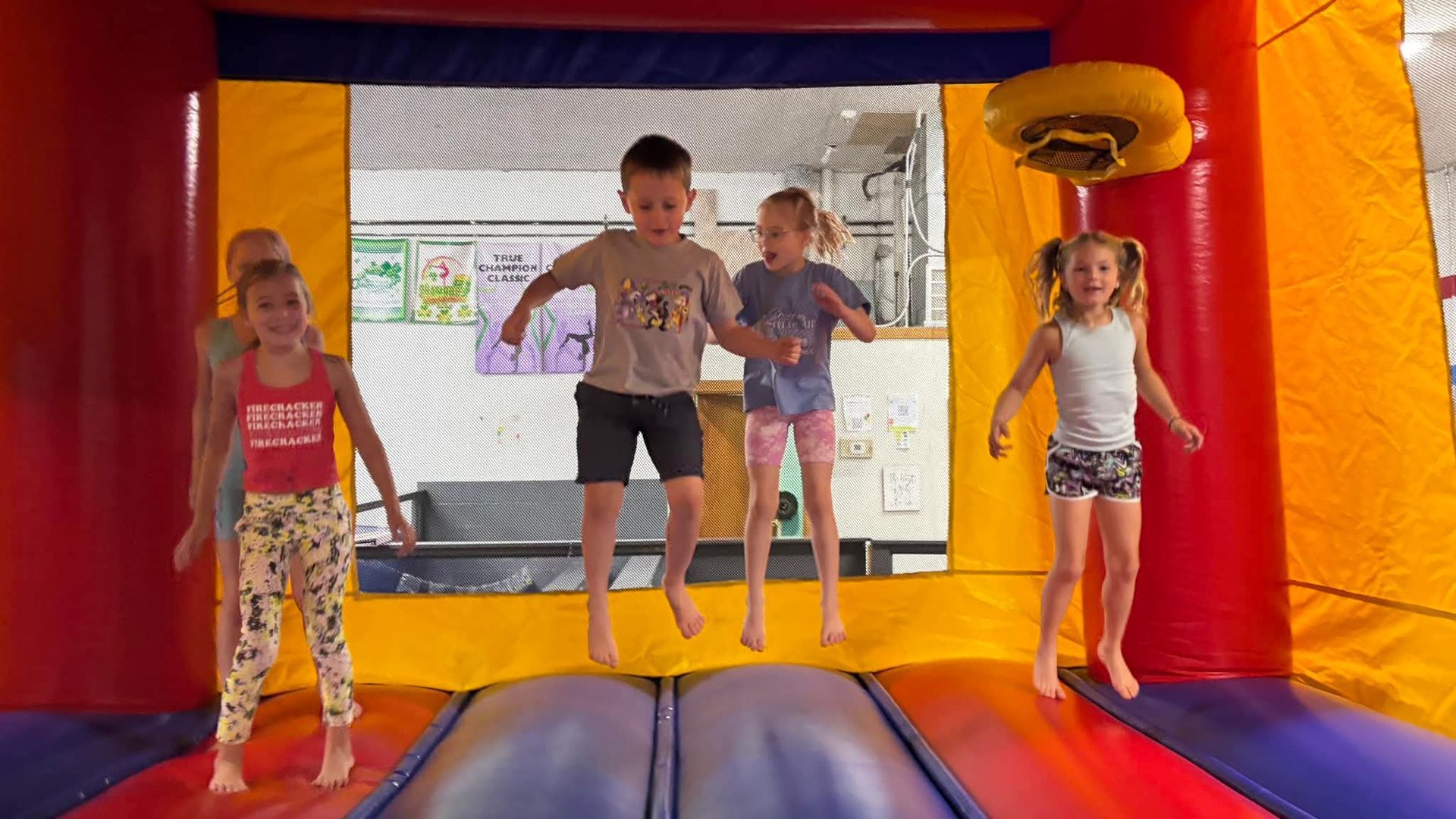 Five children jumping in a colorful inflatable bounce house with a basketball hoop.