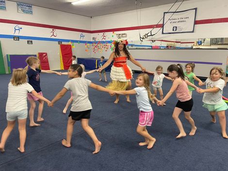 Children holding hands in a circle with an instructor in a costume; inside a gym with blue floor.