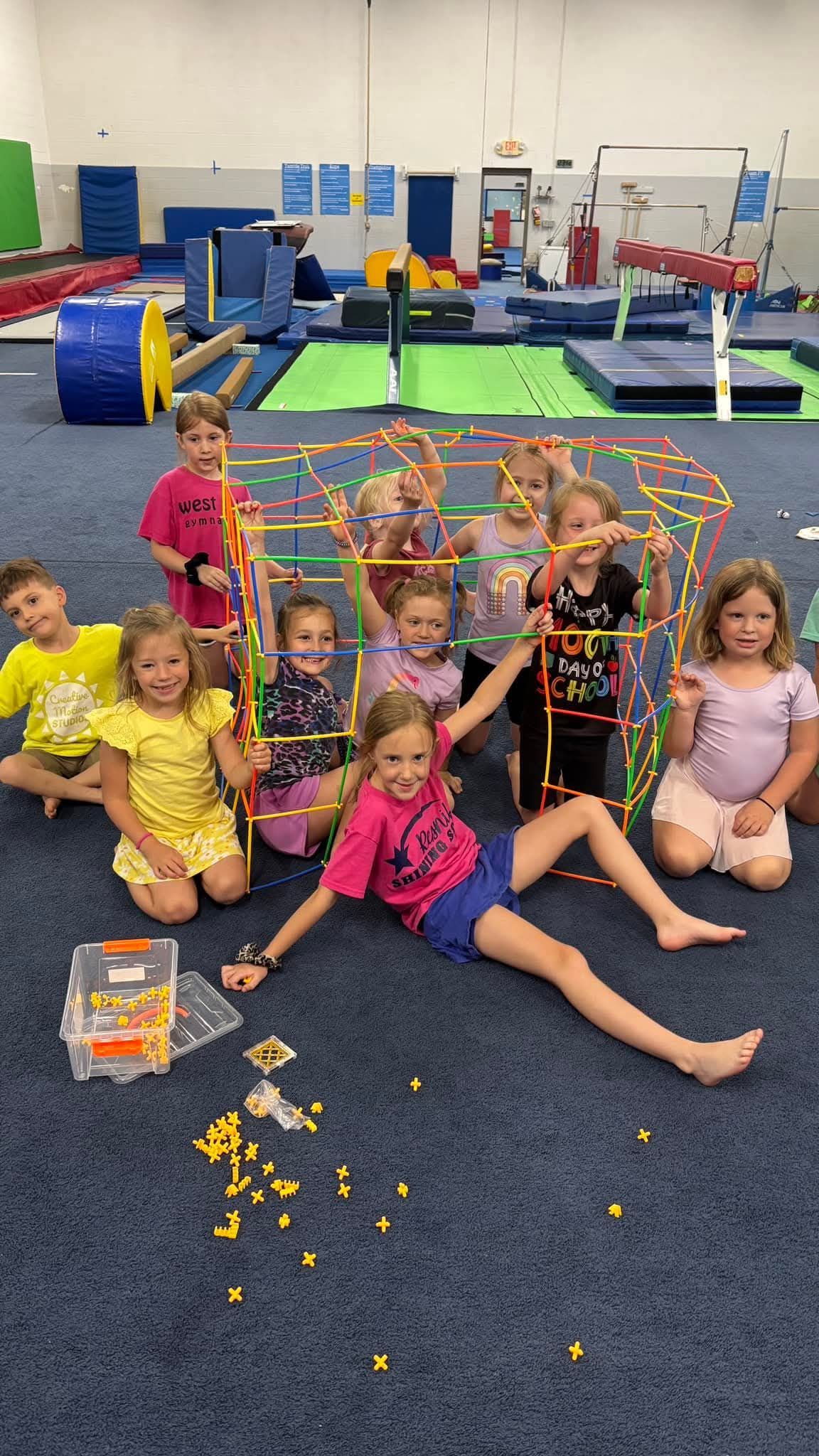 Children in a gym pose with a colorful structure made of toy pieces, smiling and waving.
