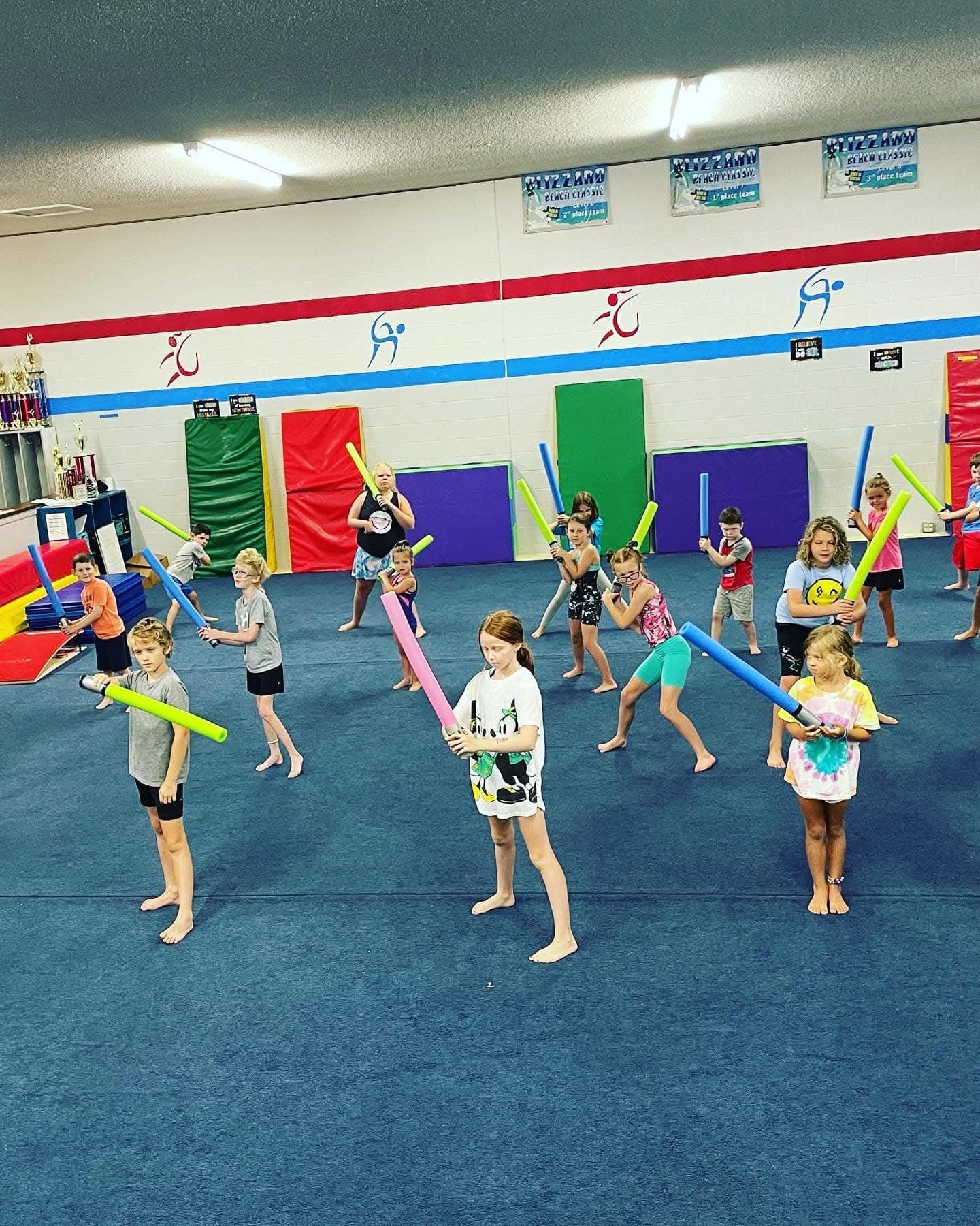 Children in a gym holding pool noodles like lightsabers. Blue floor, colorful wall decorations, white ceiling.
