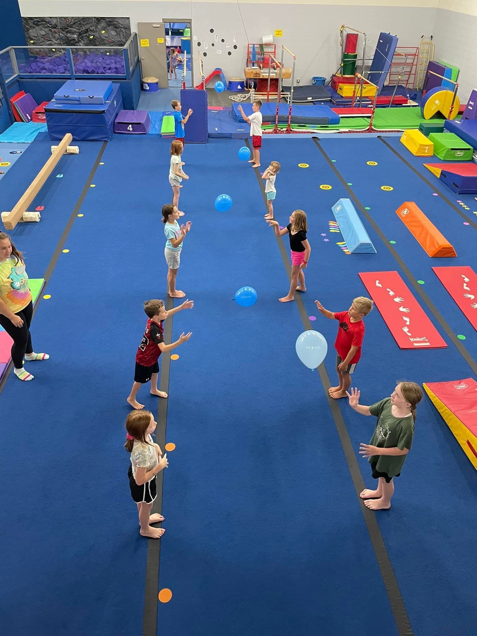 Children in a gymnastics gym toss blue balloons. Others watch. Blue mats fill the floor.