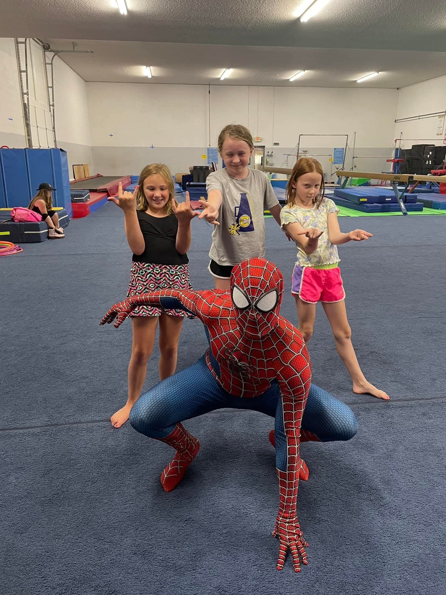 Spider-Man poses with three young girls in a gymnastics gym, all mimicking a web-shooting gesture.