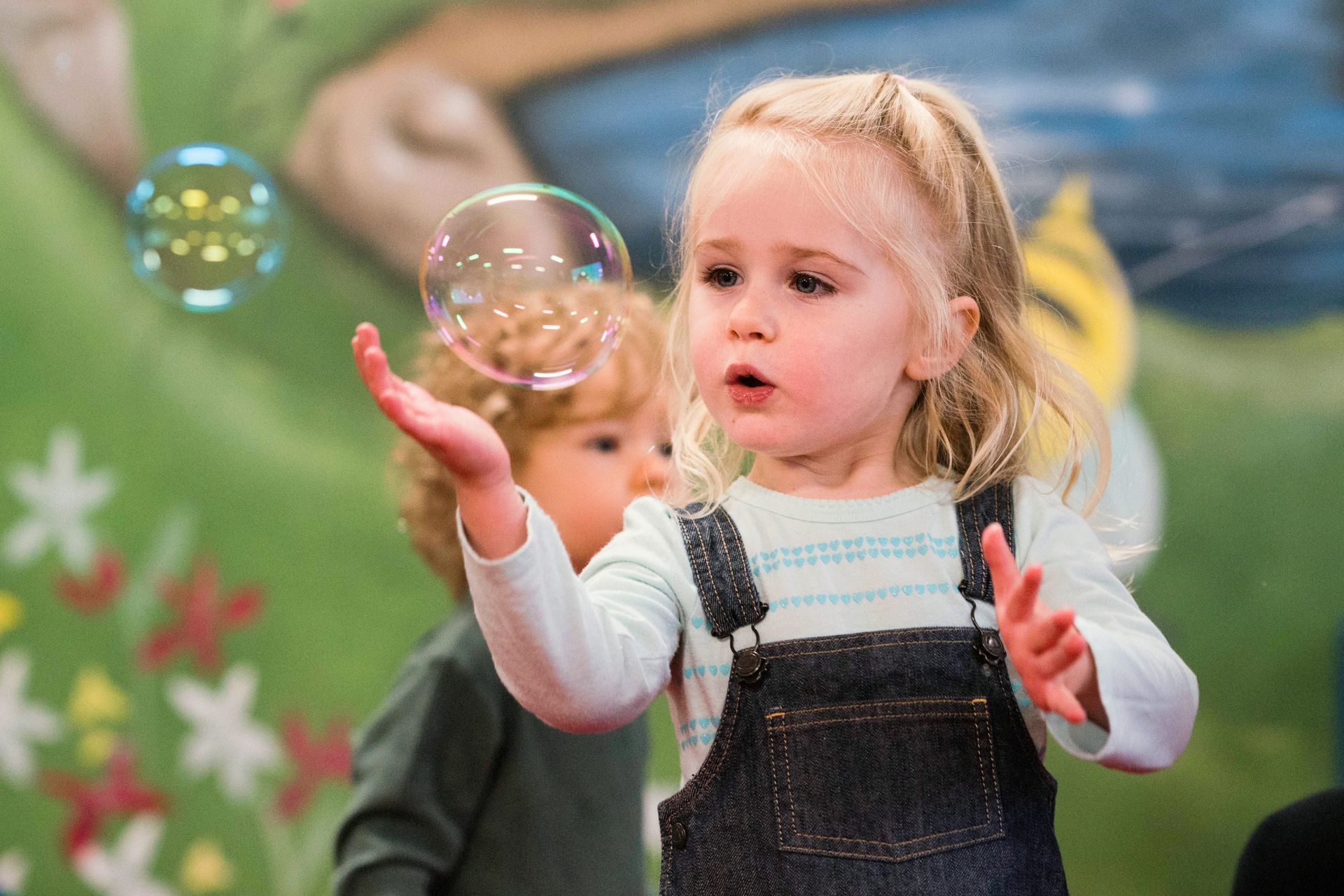 Girl in denim overalls reaches for a bubble, another child watches. Indoor setting, colorful background.