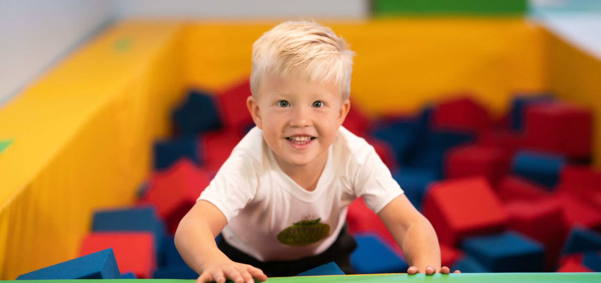Blonde child smiles, crawling on a green surface toward the viewer in a pit filled with foam cubes.