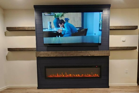 Wall-mounted TV above a modern electric fireplace with rustic wood shelves on a beige wall.