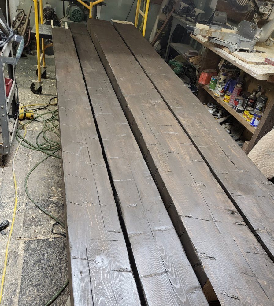 Long wooden planks laid side by side in a cluttered workshop or garage, with tools and shelves nearby
