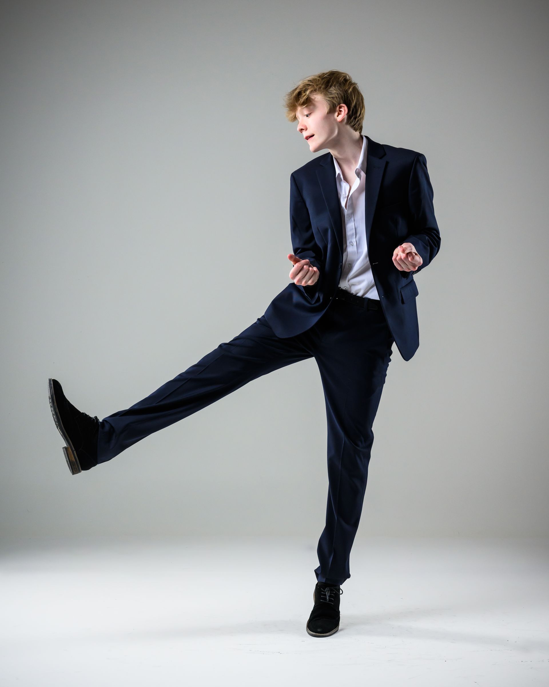 Person in a navy suit kicking one leg up against a gray studio background.
