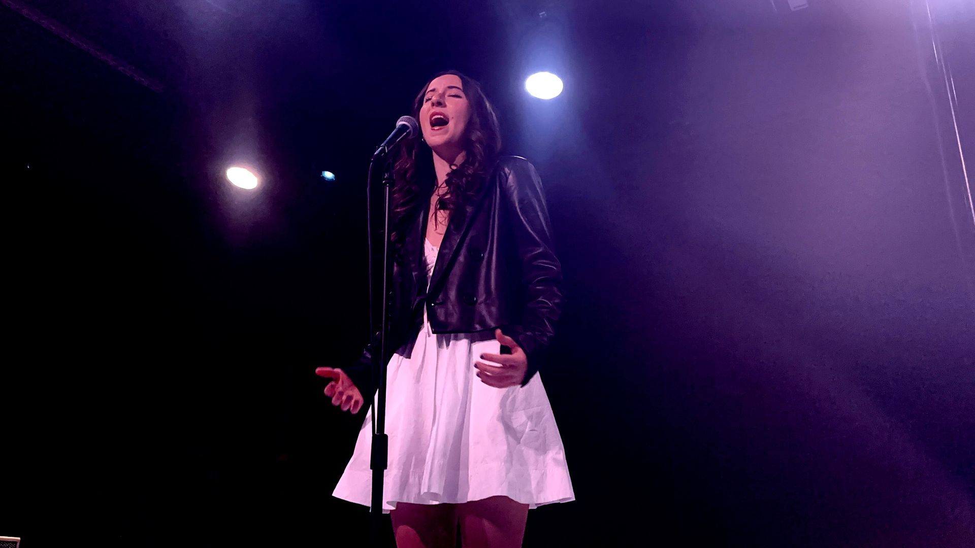 Singer performing onstage under purple lights, wearing a black jacket and white skirt, singing into a microphone.
