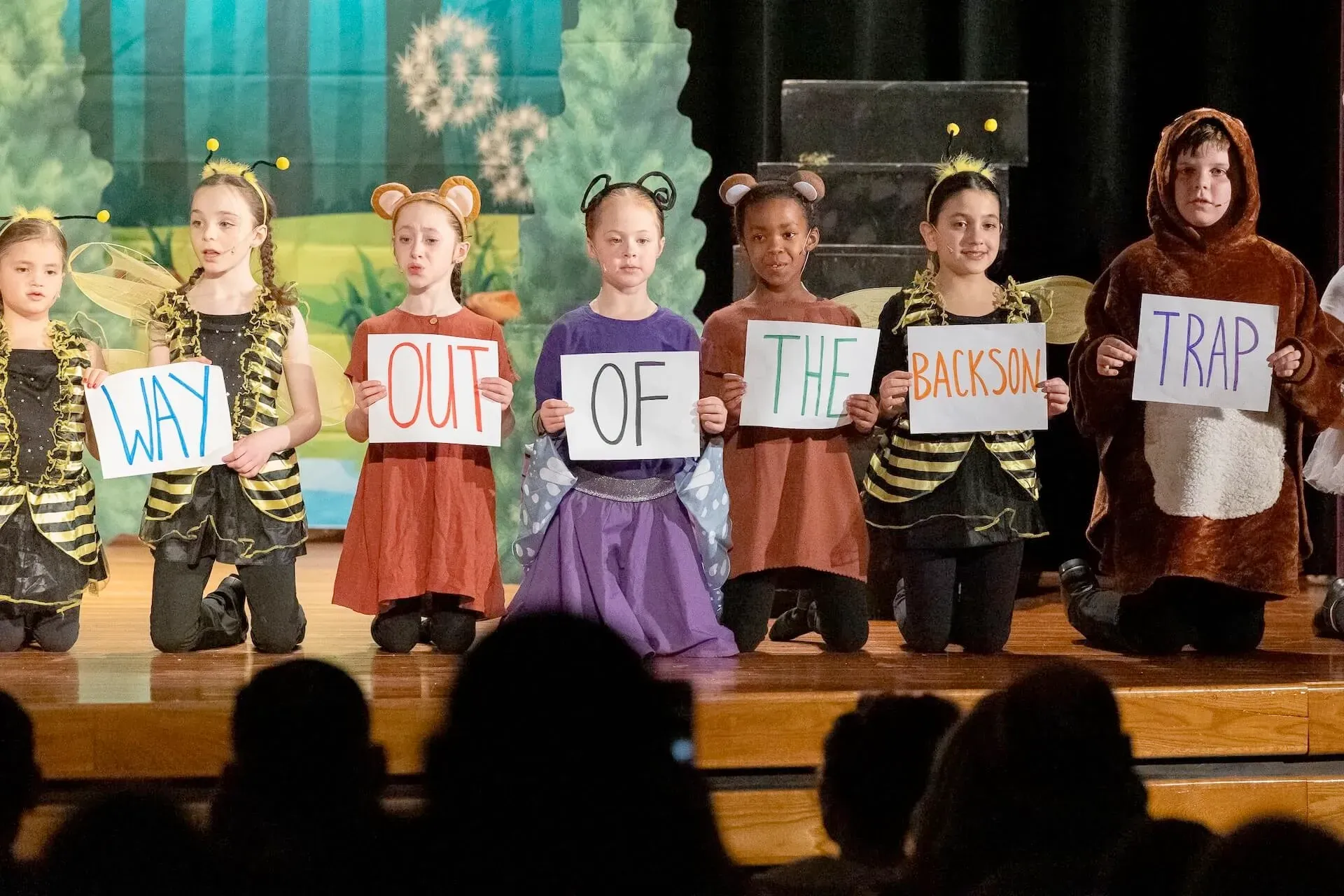 Children on stage holding signs spelling “WHAT IS OUT OF THE BOX TRAP?”