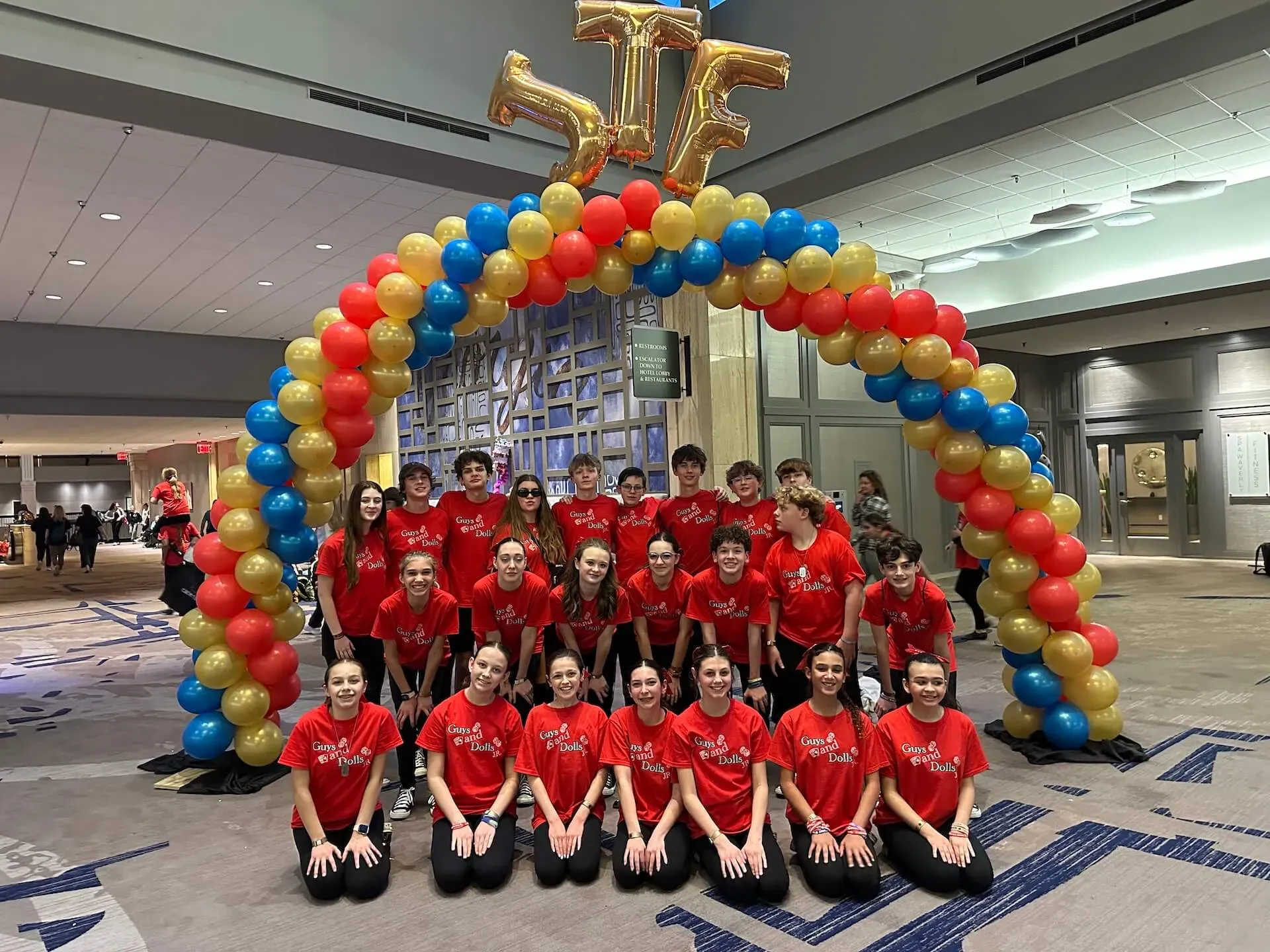 Group in red shirts posing under a red, blue, and yellow balloon arch in a convention hall.