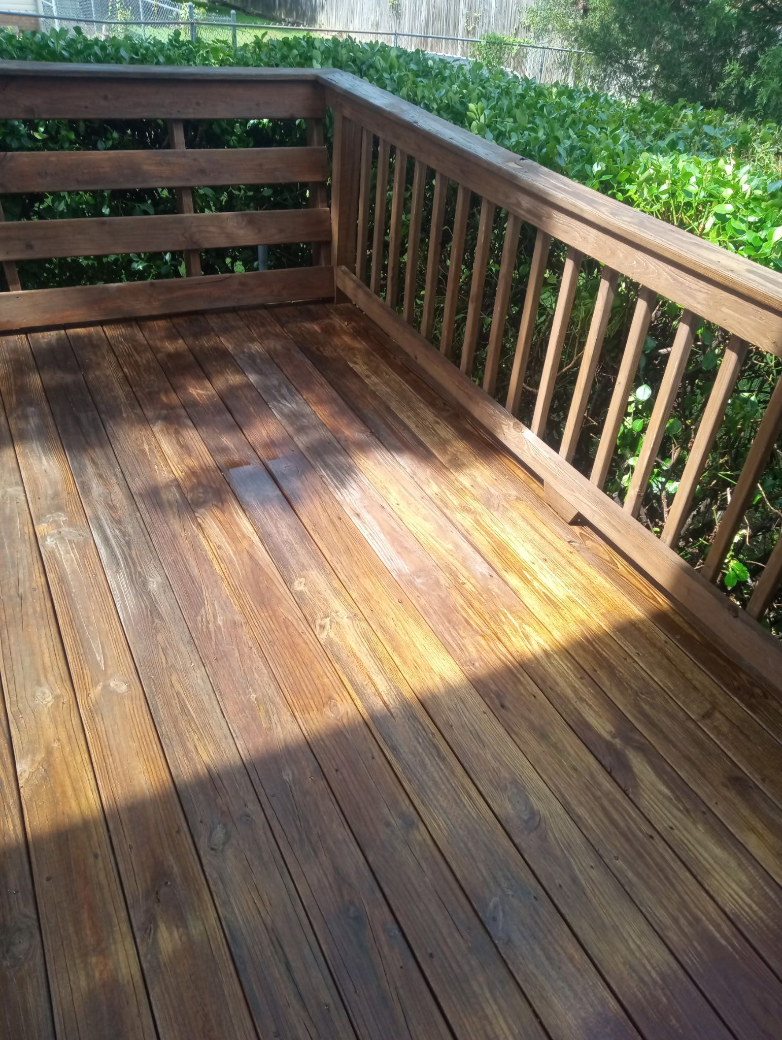 Wooden deck with brown stain, partially weathered, surrounded by a railing and greenery.