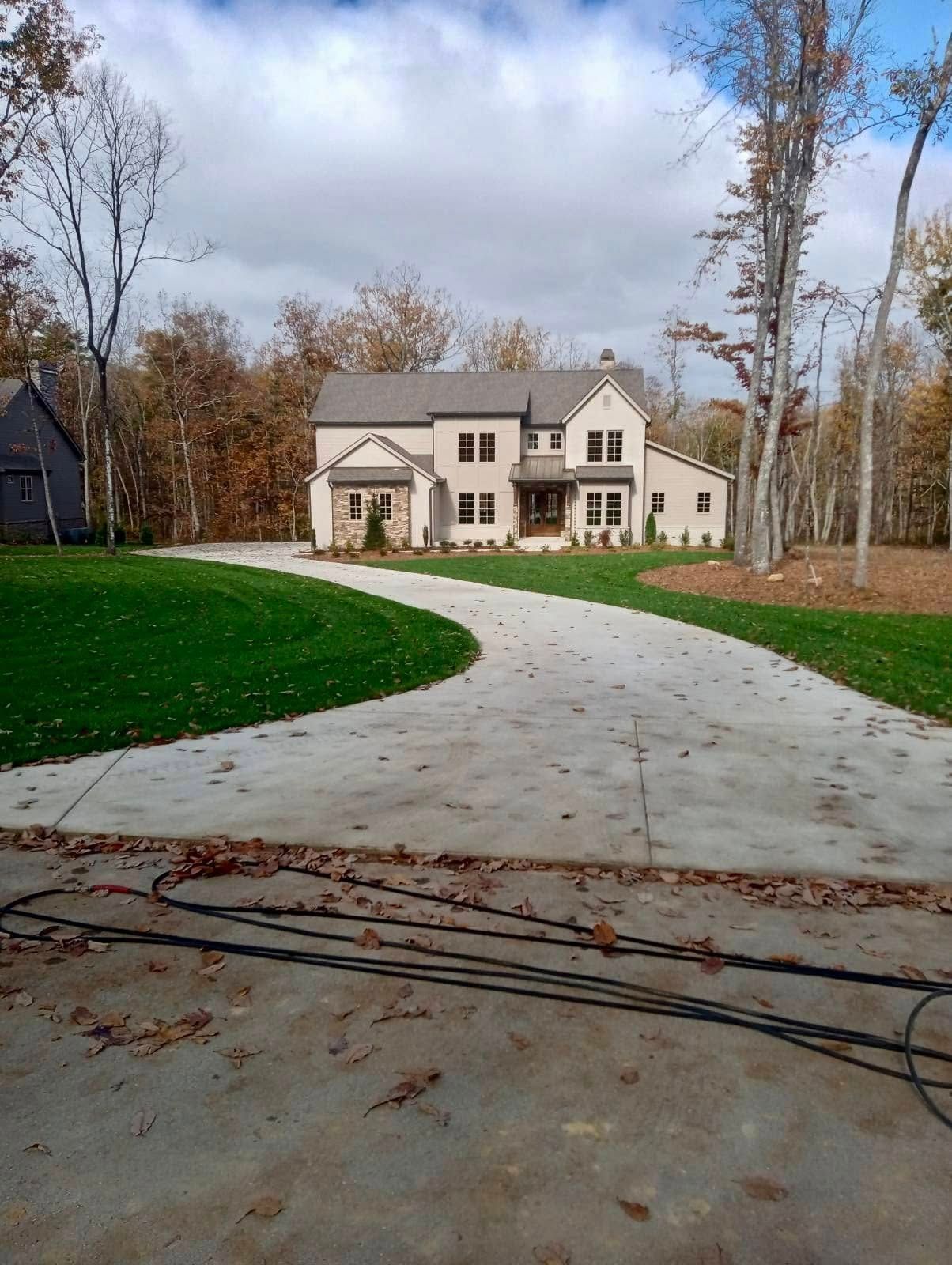 A two-story beige house with a winding driveway; green lawn in front, fall foliage behind.