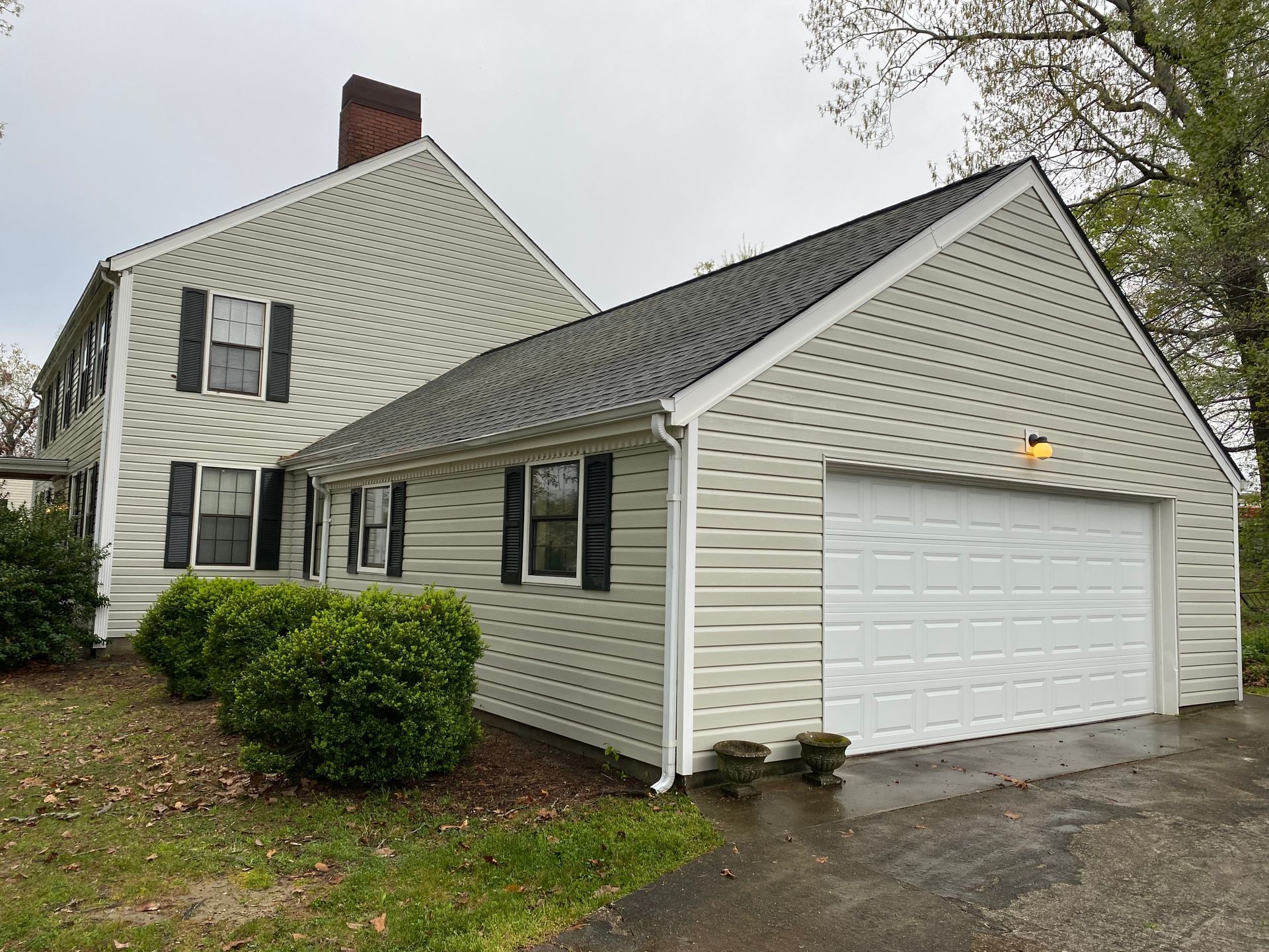 Two-story house with attached garage. Light beige siding, black shutters, green bushes, white garage door, and gray roof.
