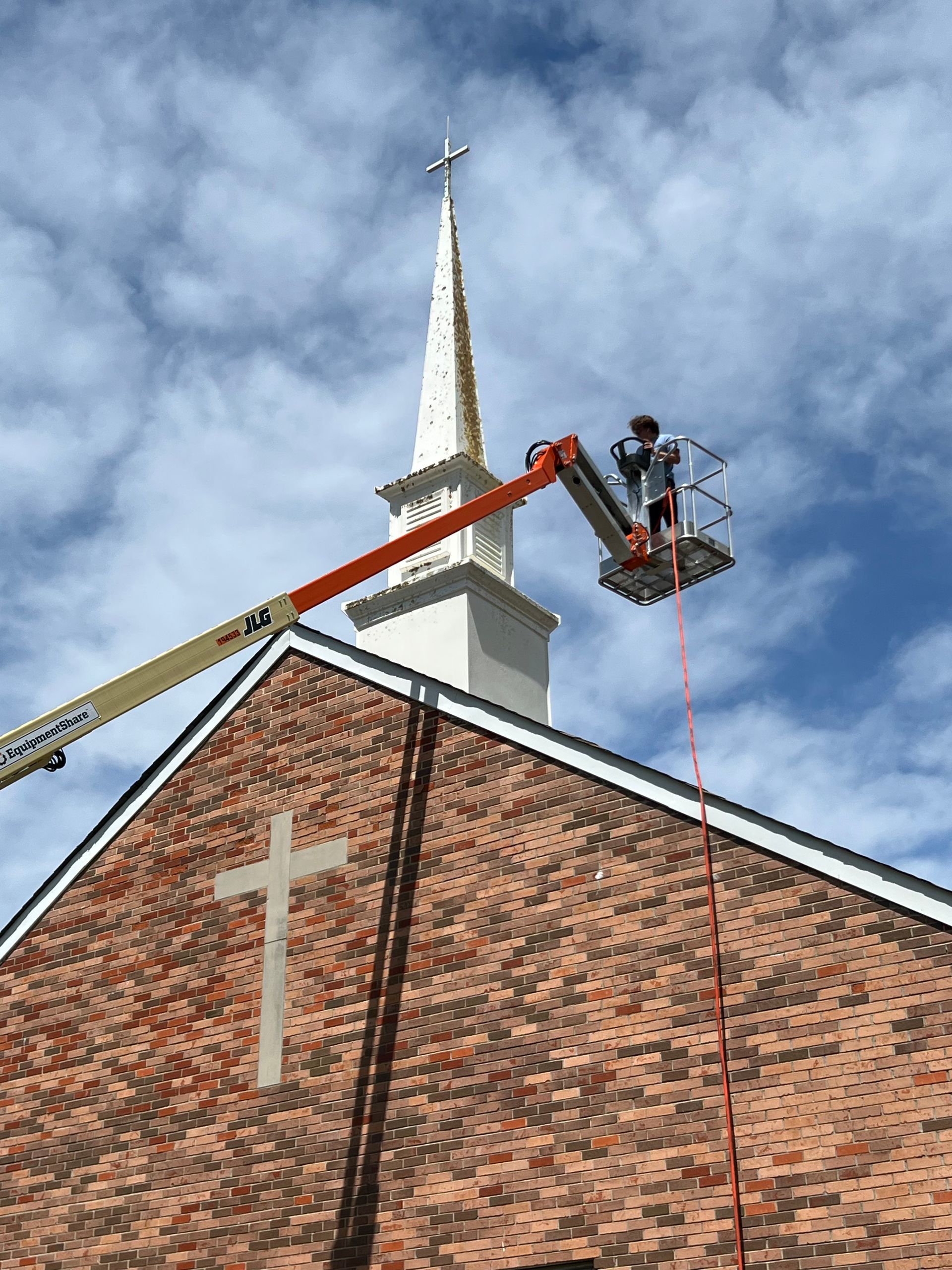 Workers in a lift repairing a church steeple above a red brick roof under a cloudy sky