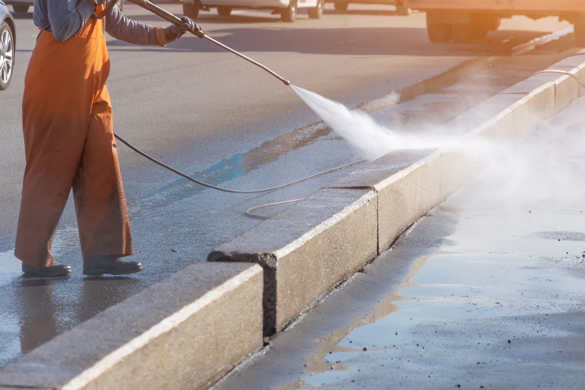 Person in orange overalls power washing a concrete curb on a city street; water sprays.