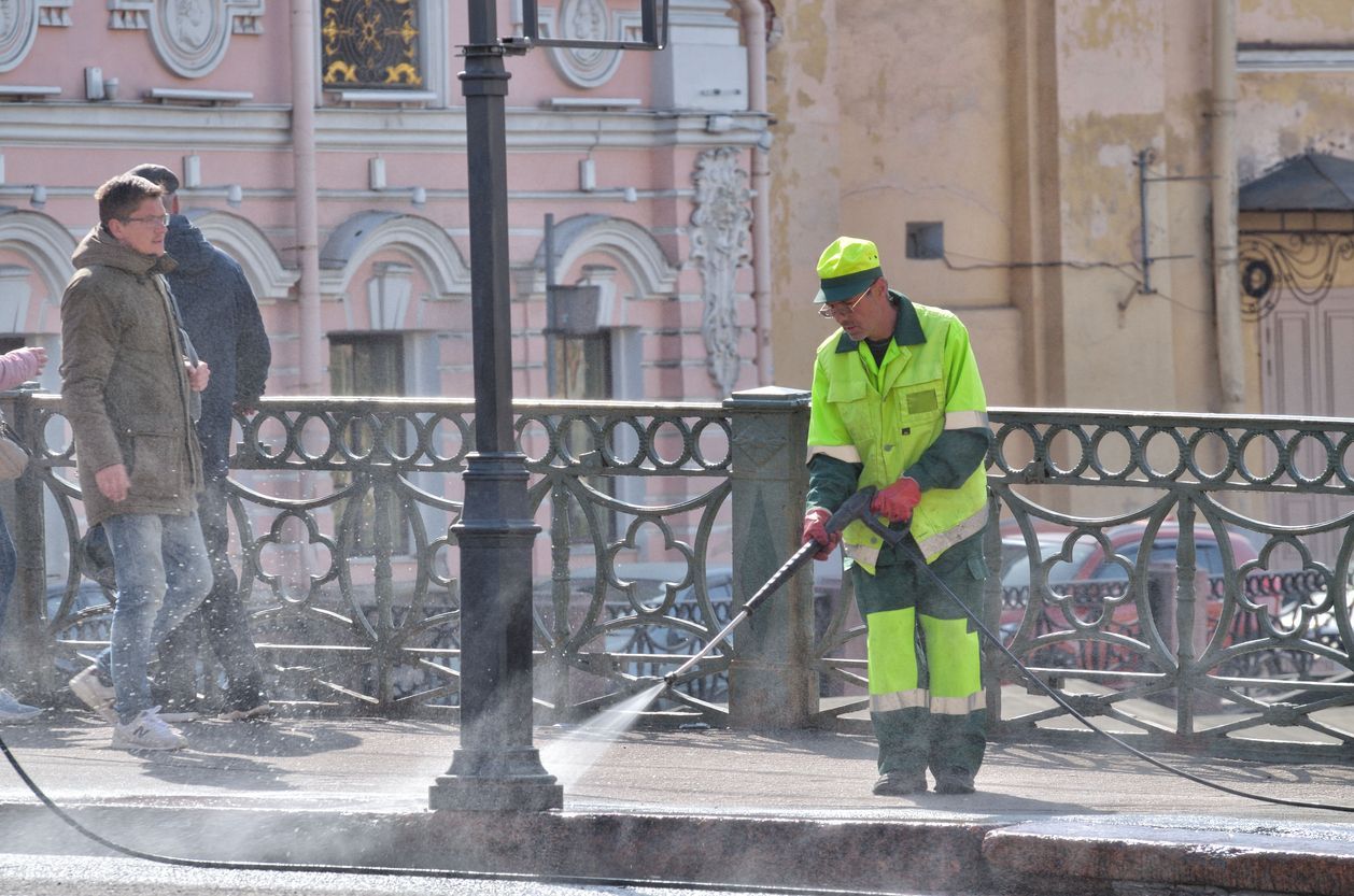 A sanitation worker in neon green scrubs cleans a stone walkway near a black iron fence and onlookers.