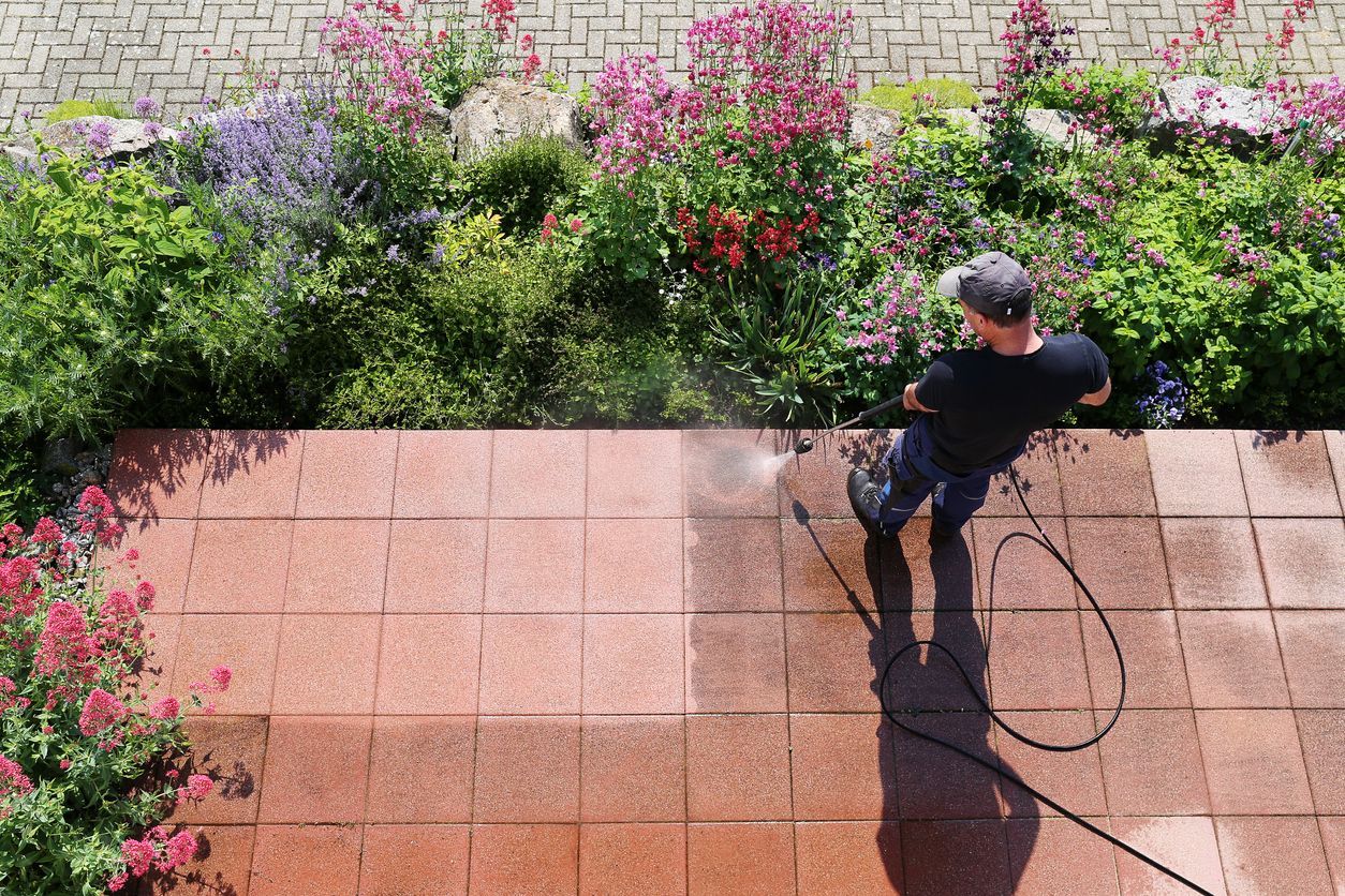 Person power washing a red brick patio next to a garden of flowers.